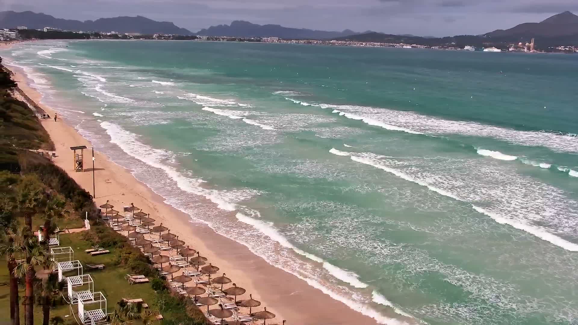 A tranquil, clear turquoise bay with a sandy beach in the foreground is visible under a partly cloudy sky, with a distant town and mountainous coastline across the water.