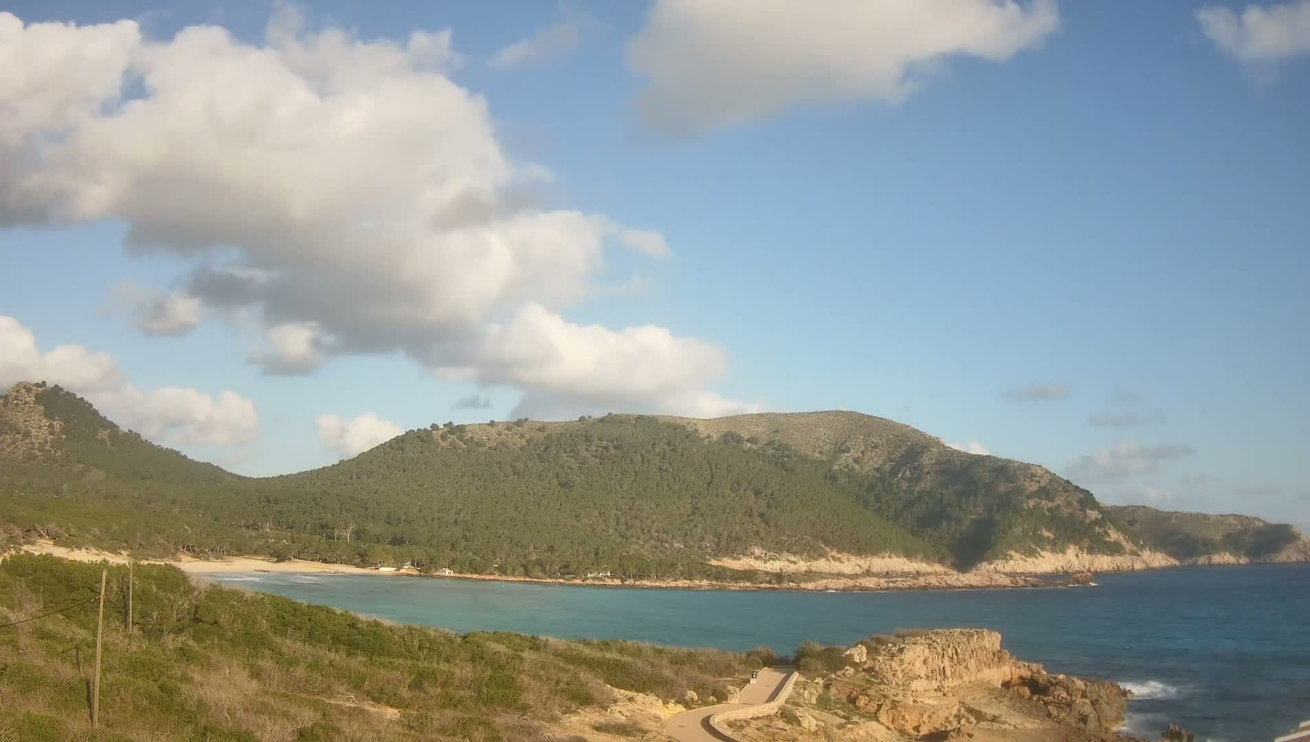 A partly cloudy sky overlooks a secluded cove with a small sandy beach, surrounded by green hills and rocky coastline.