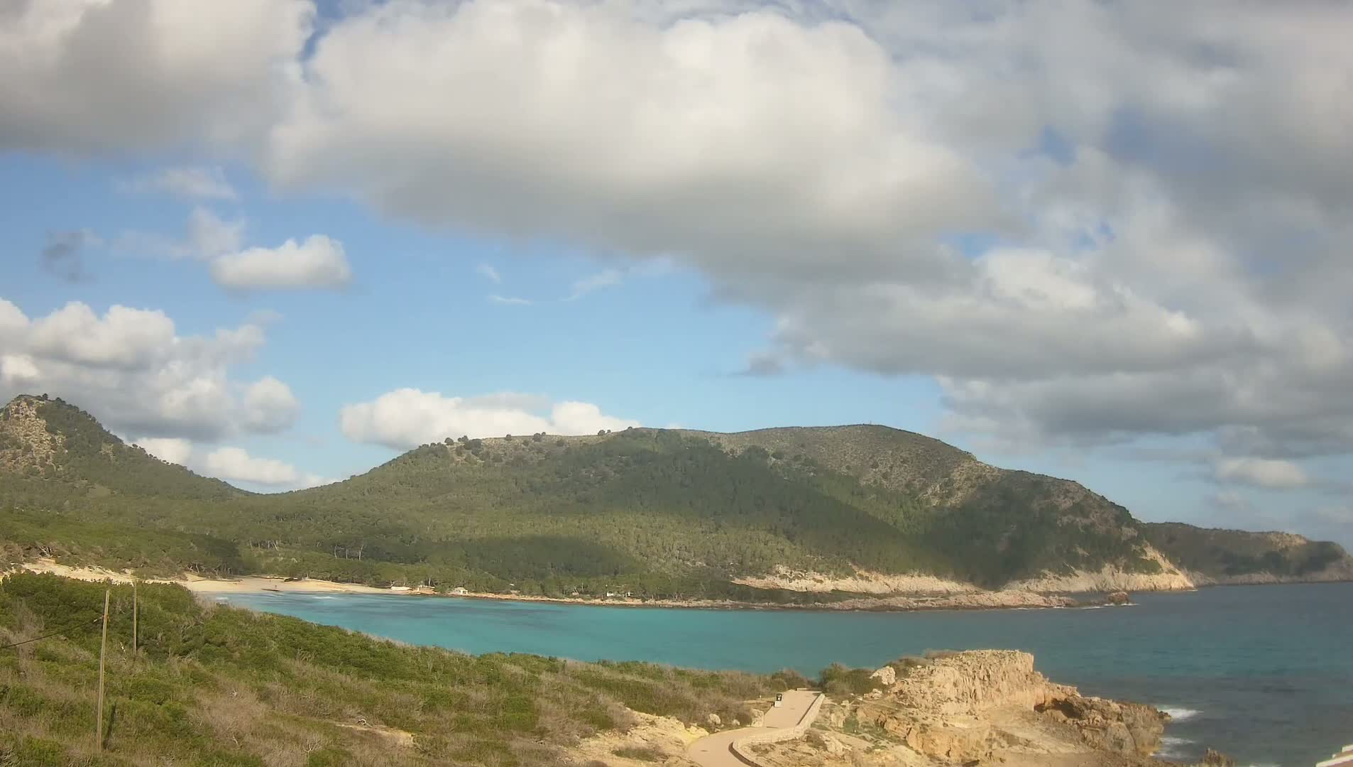 A partly cloudy day reveals a secluded cove with turquoise water, a sandy beach, and lush green hillsides under a mostly clear sky.