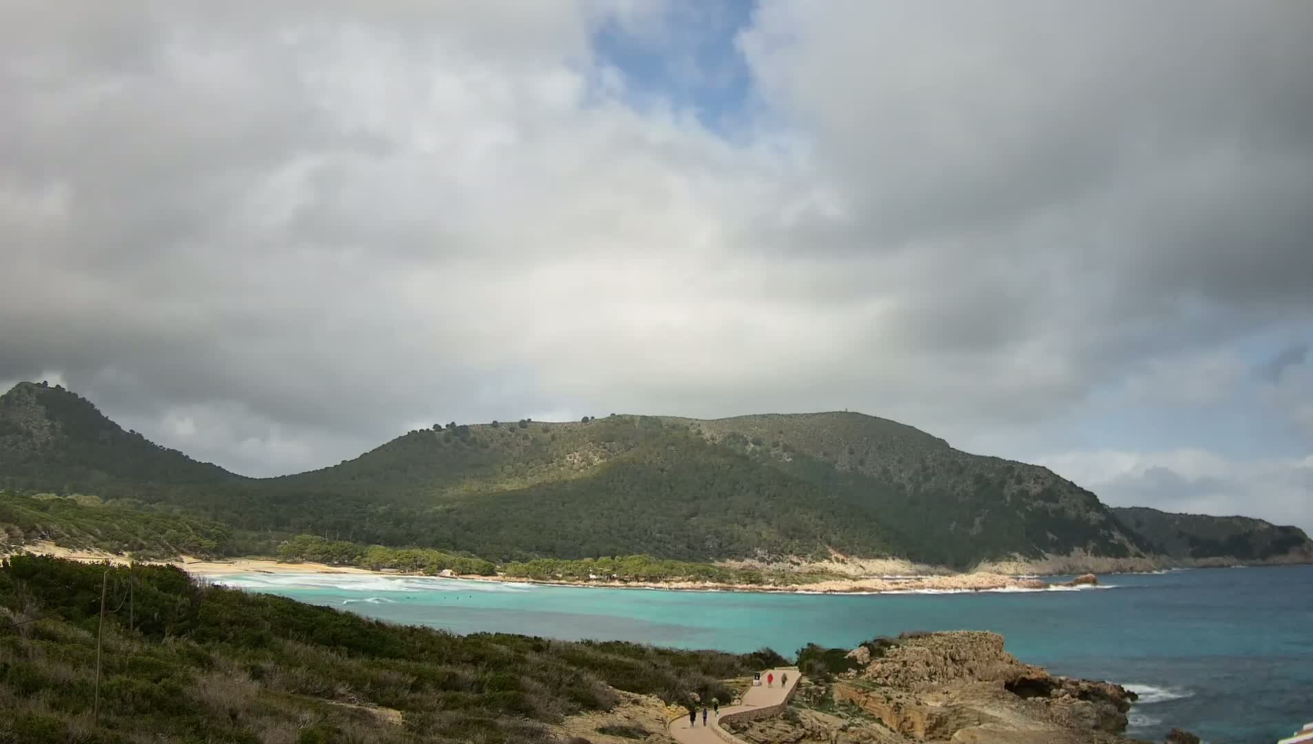 A mostly cloudy sky overlooks a turquoise bay with a sandy beach, backed by green hills and a walking path.