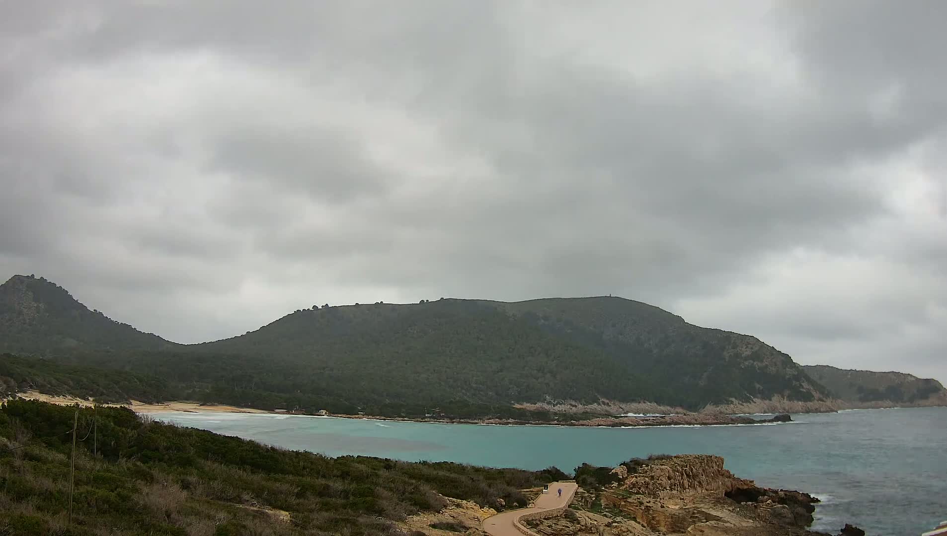 A person walks along a path overlooking a tranquil, turquoise bay nestled between two verdant hills under a cloudy sky.