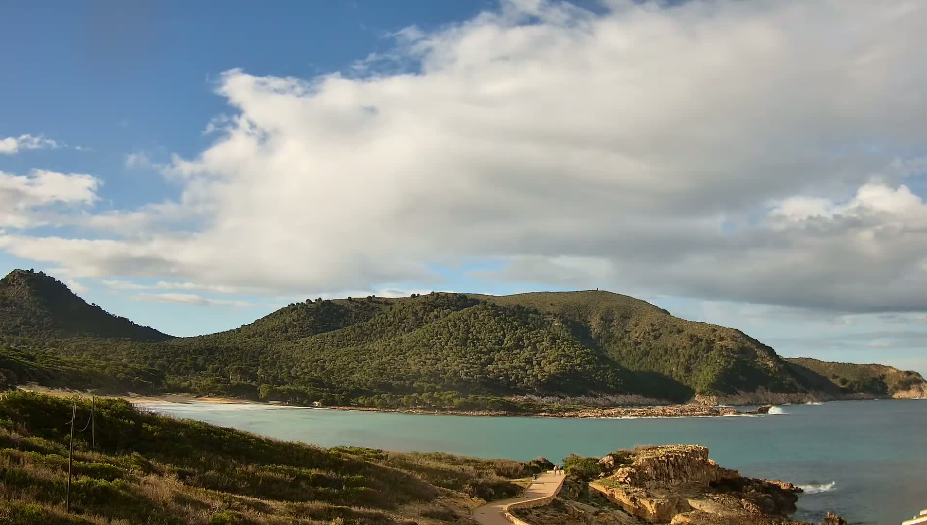 A partly cloudy day reveals a secluded cove with a sandy beach nestled between two verdant, tree-covered hills, the turquoise water gently lapping at the rocky shore.