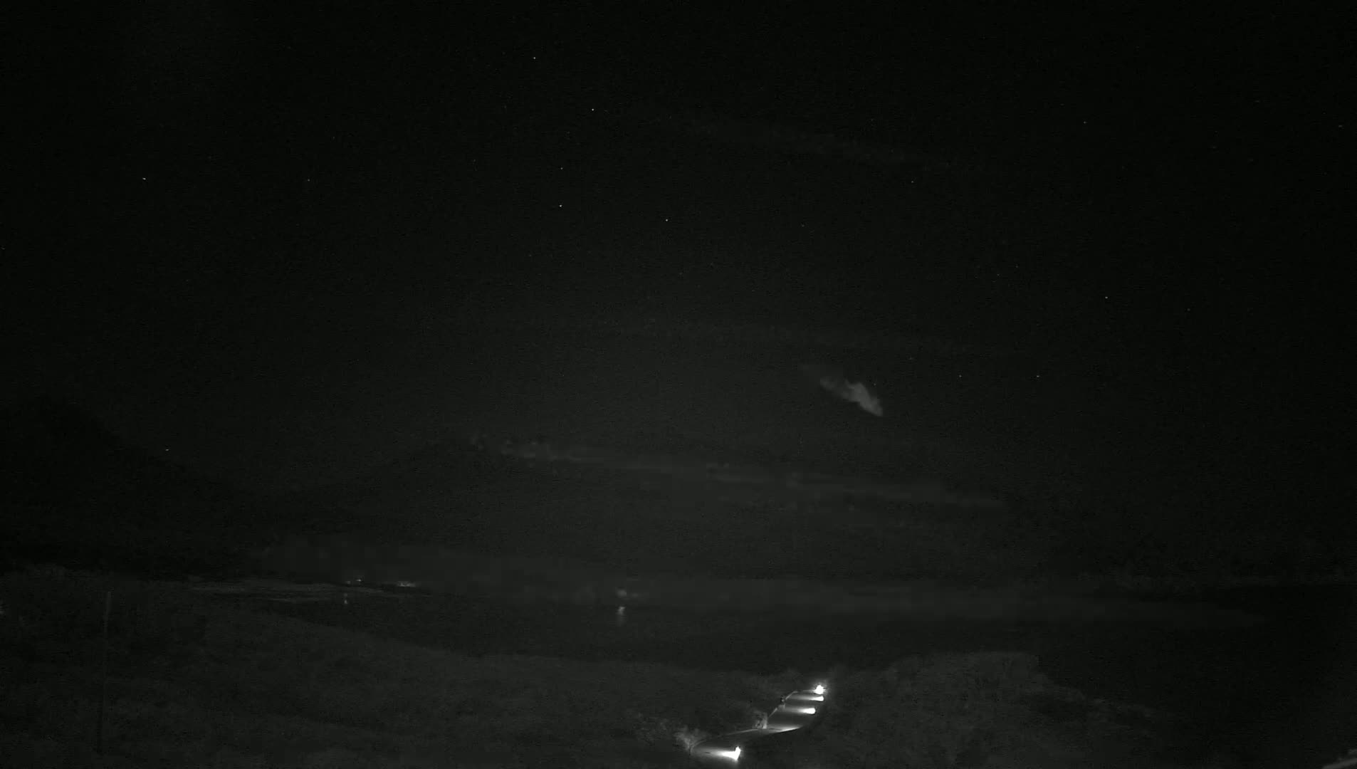 A nighttime view of a dark, cloudy sky over a calm body of water, with a road and faint lights visible in the foreground.