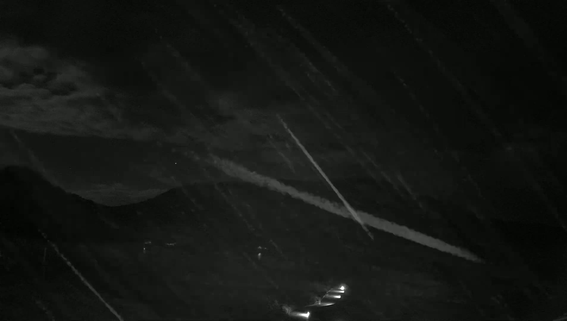 At night, streaks of light appear in a dark sky over dark mountains, with light rain or snow falling.