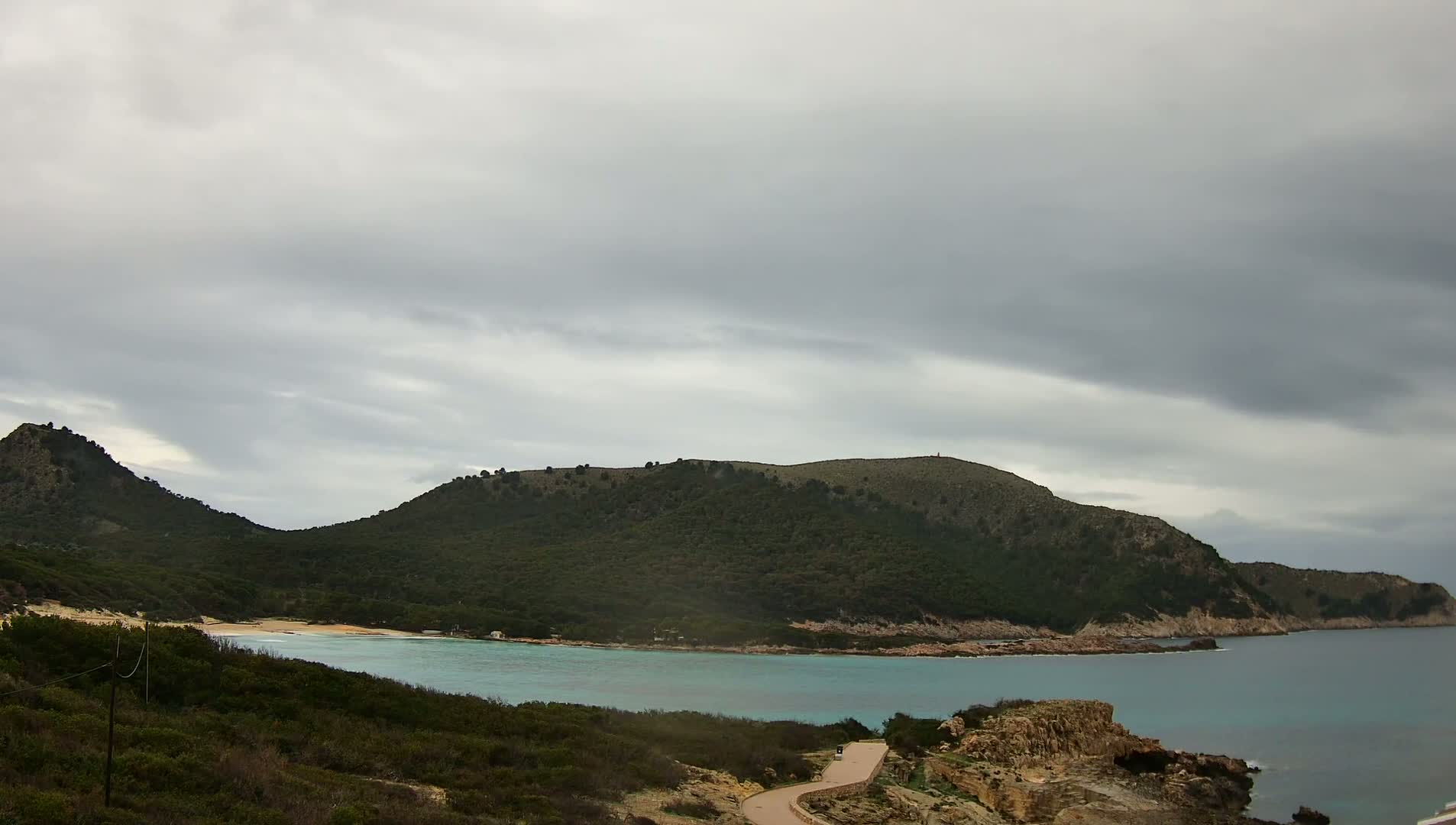 A secluded cove with turquoise water is nestled between two verdant hills under a cloudy sky.