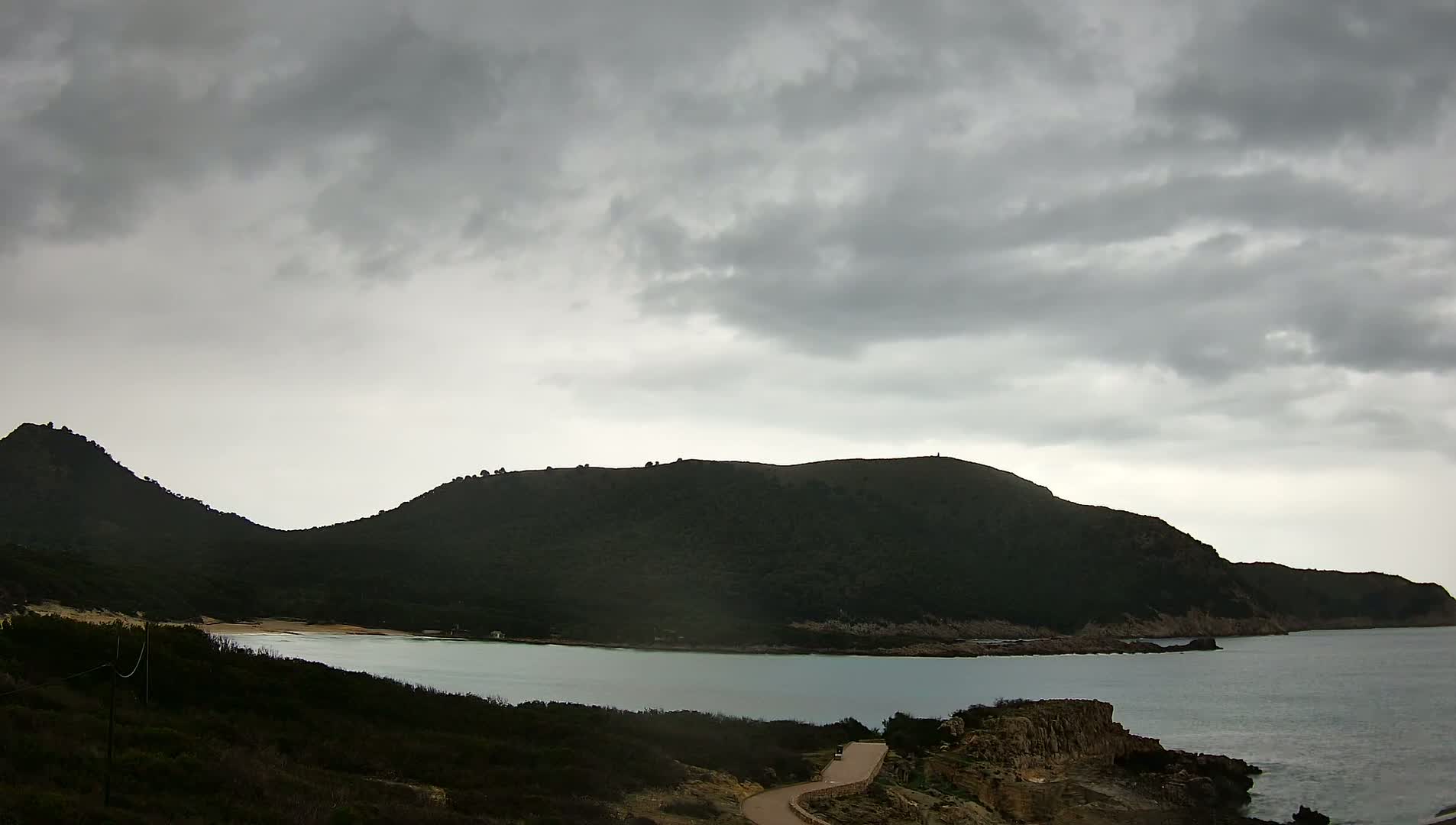 A winding road leads down to a calm bay nestled between dark, verdant hills under a cloudy sky.