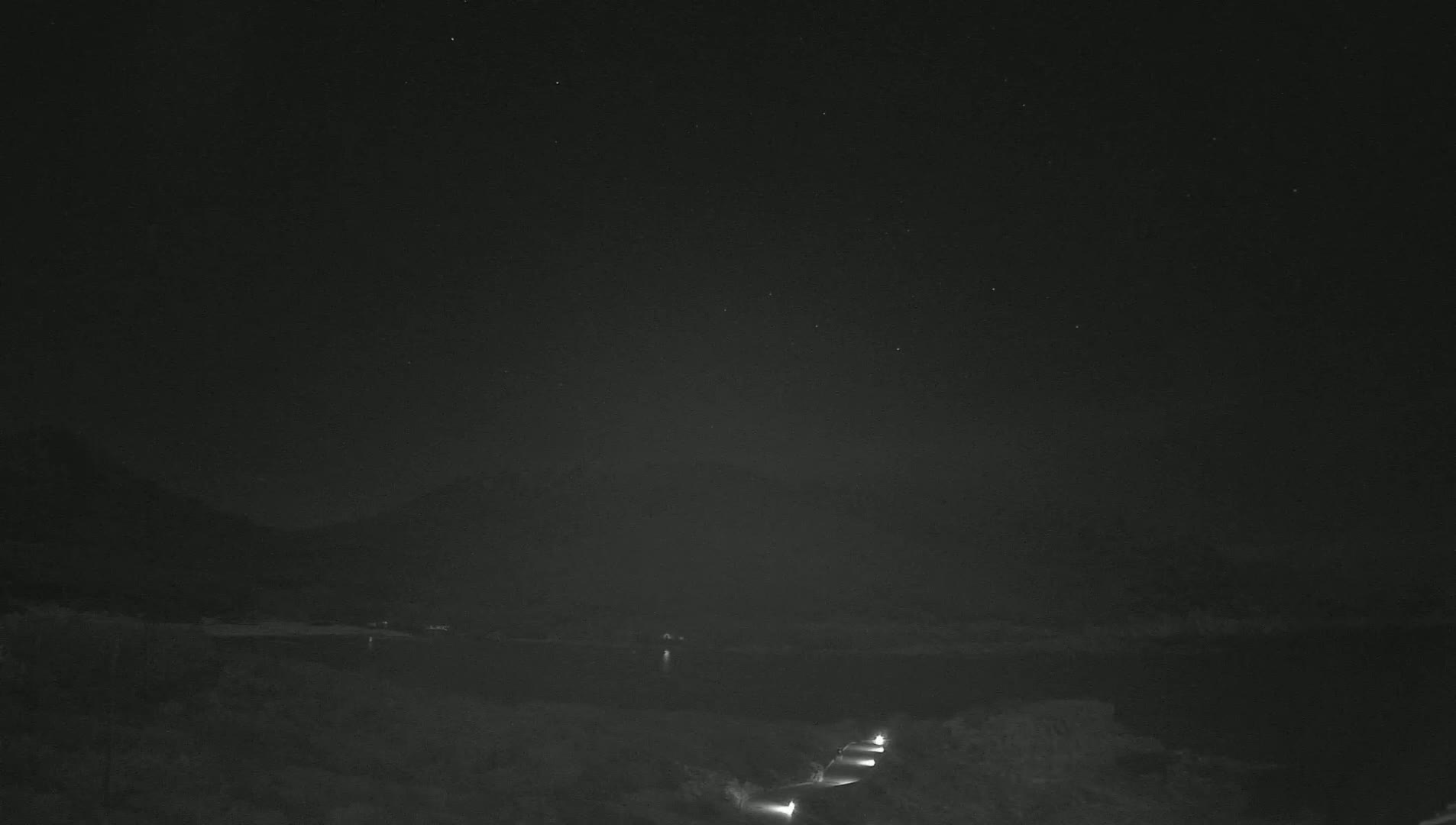 A nighttime, low-light view shows a dark body of water under a clear, starry sky, with a road and some lights visible in the foreground.