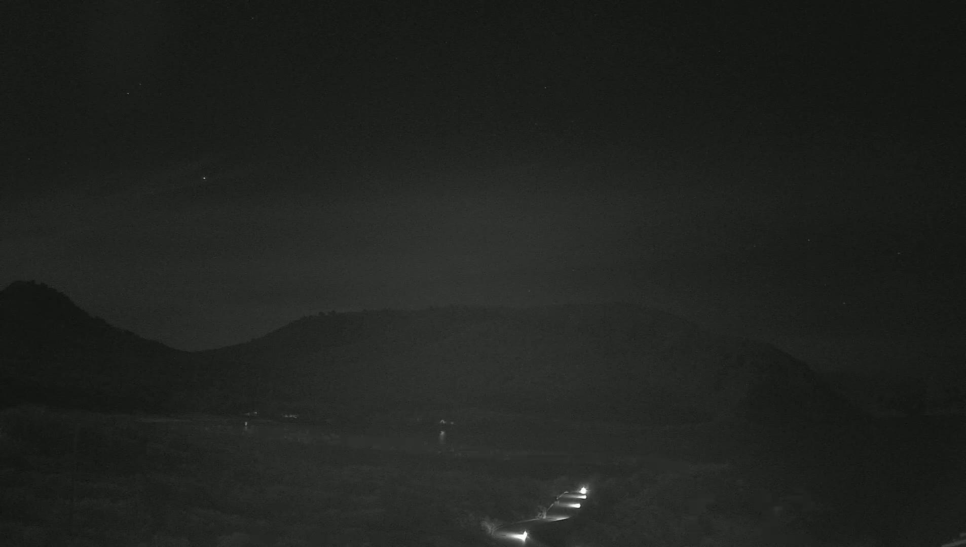 Under a clear, dark night sky, headlights from several vehicles are visible on a road winding through a low mountain pass.