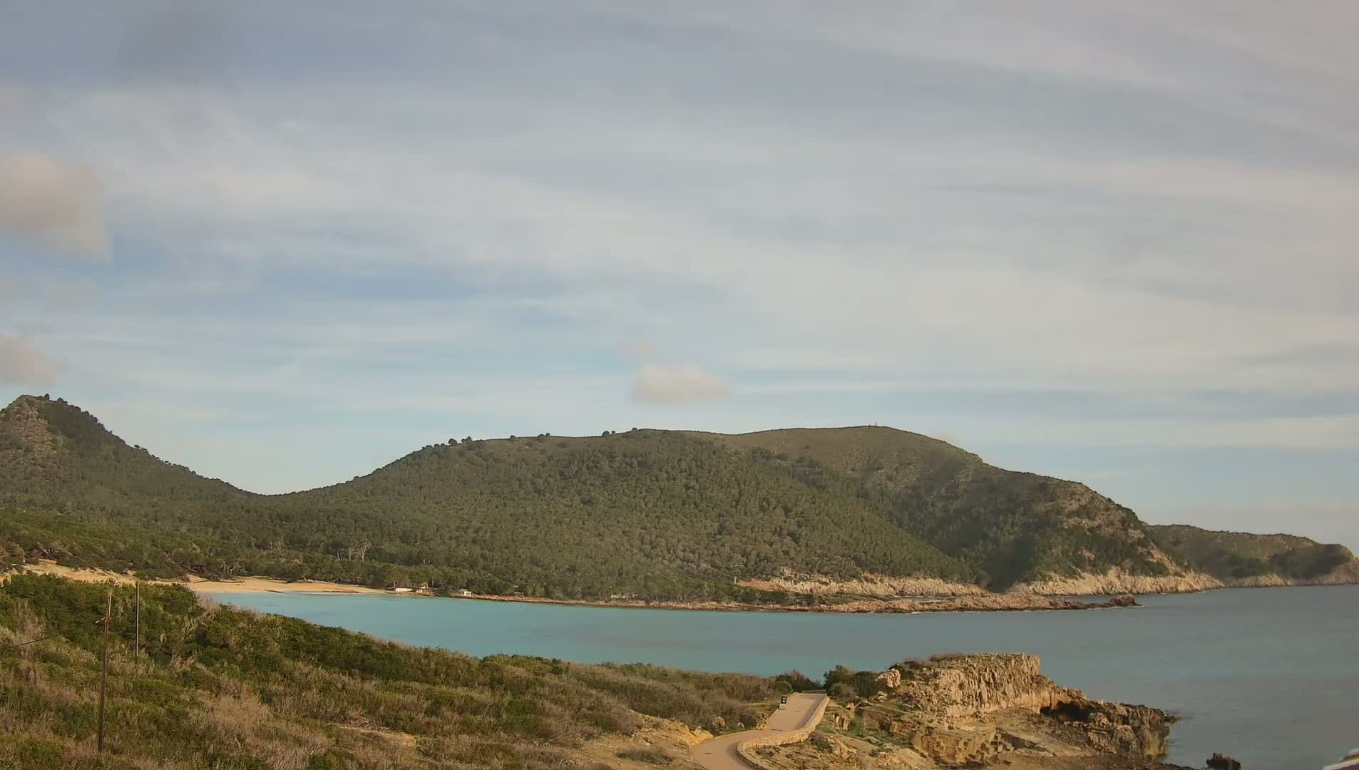 A tranquil cove with a small sandy beach nestled between two verdant hills under a partly cloudy sky.