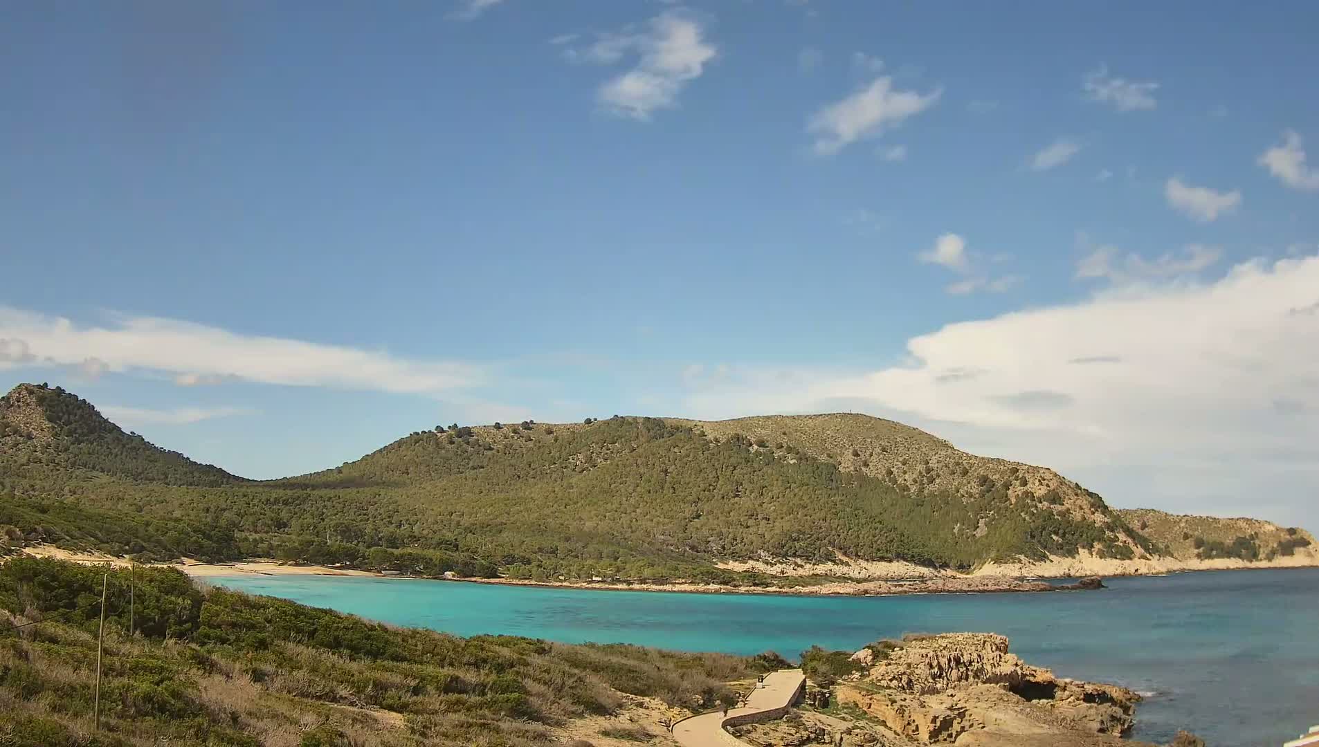 A secluded cove with turquoise water is nestled between two verdant hills under a mostly sunny sky with scattered clouds.