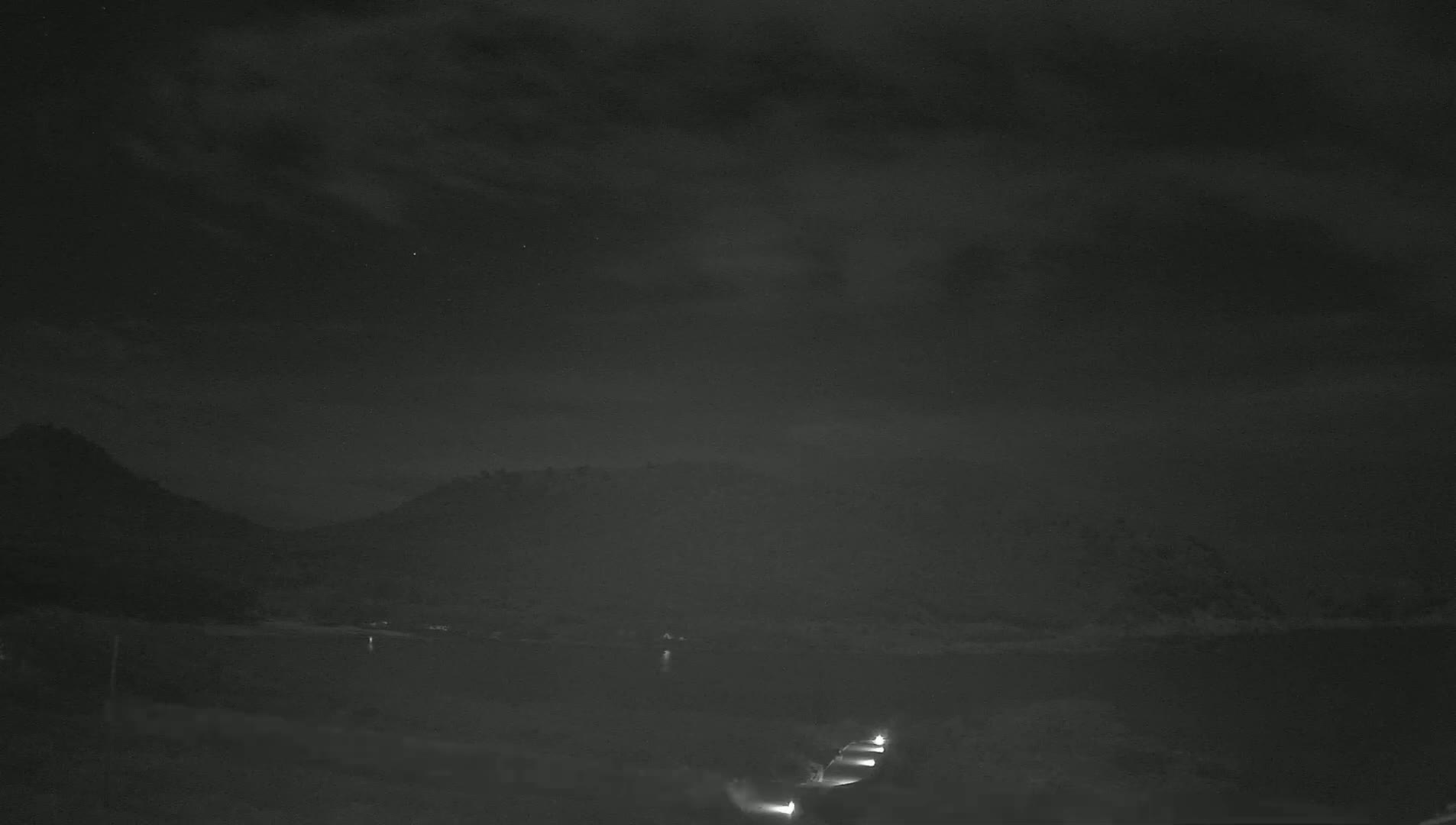 A nighttime scene shows a dark, calm body of water reflecting a cloudy sky, with dimly lit hills in the background and faint lights visible on a structure near the water's edge.