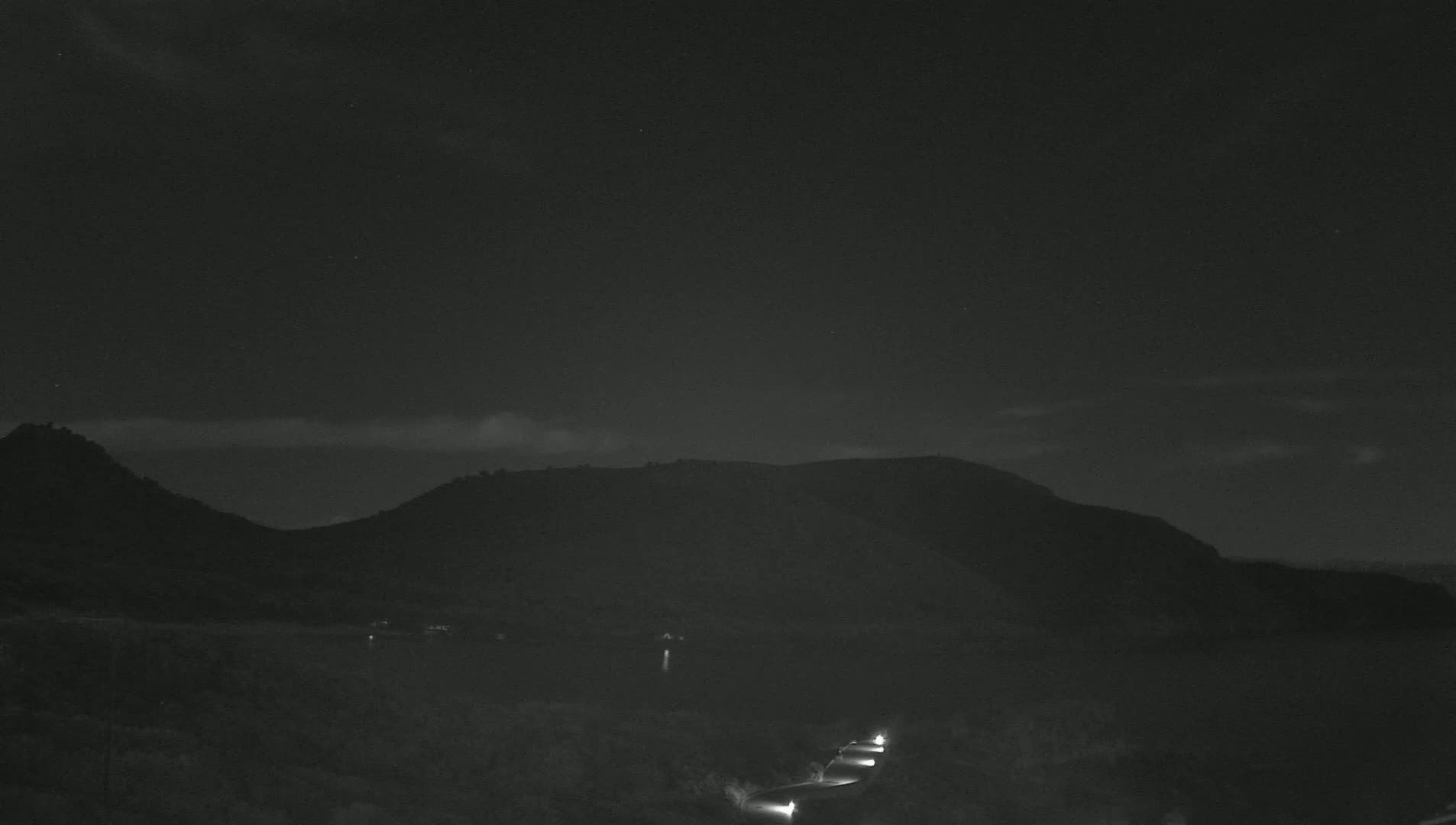 Headlights from several vehicles are visible on a road at night, with dark mountains and a mostly clear sky in the background.