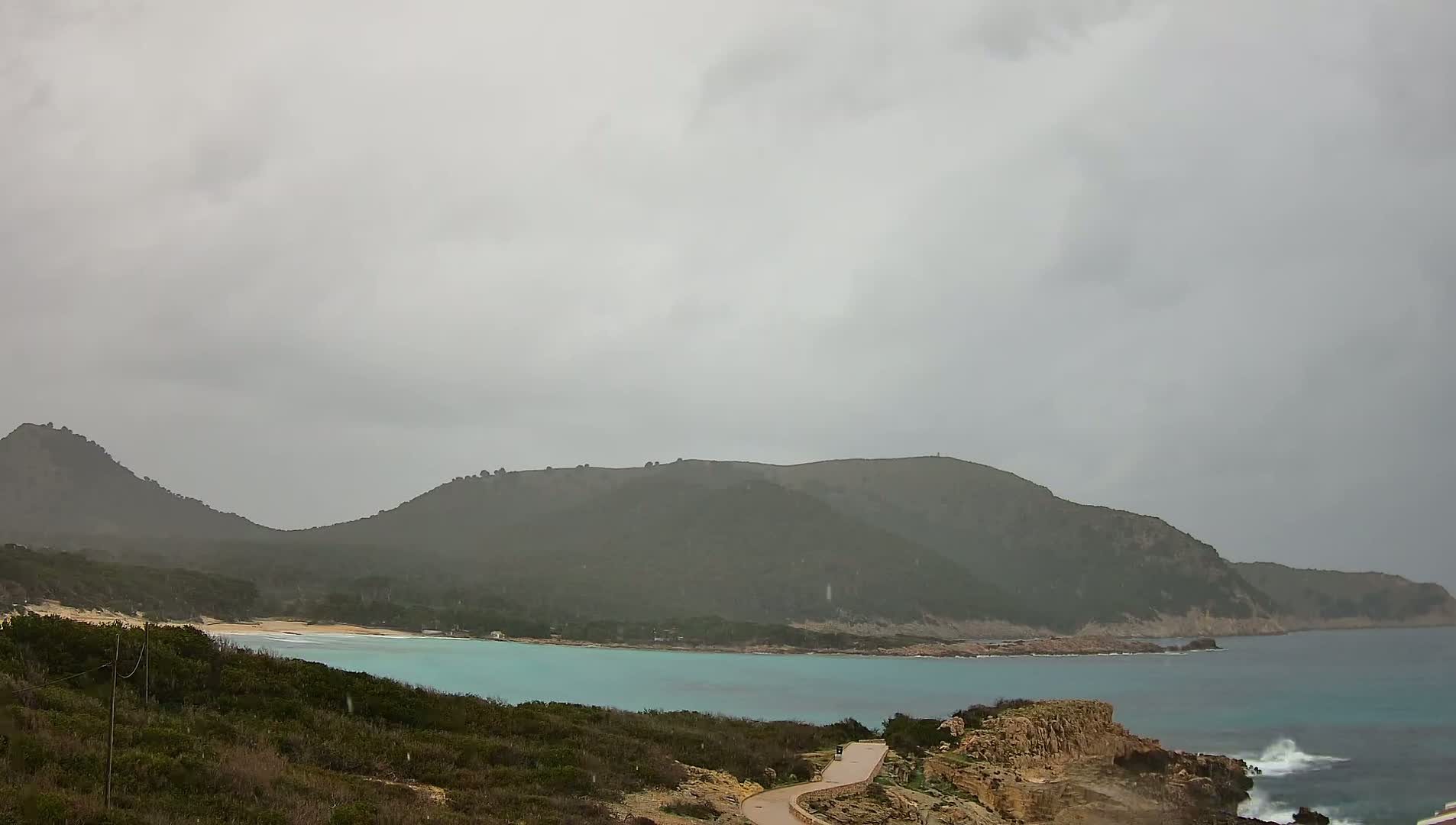 A coastal scene under an overcast sky shows a tranquil, turquoise bay nestled between dark green hills, with a light-colored sandy beach visible in the distance and some light wave action near the foreground.