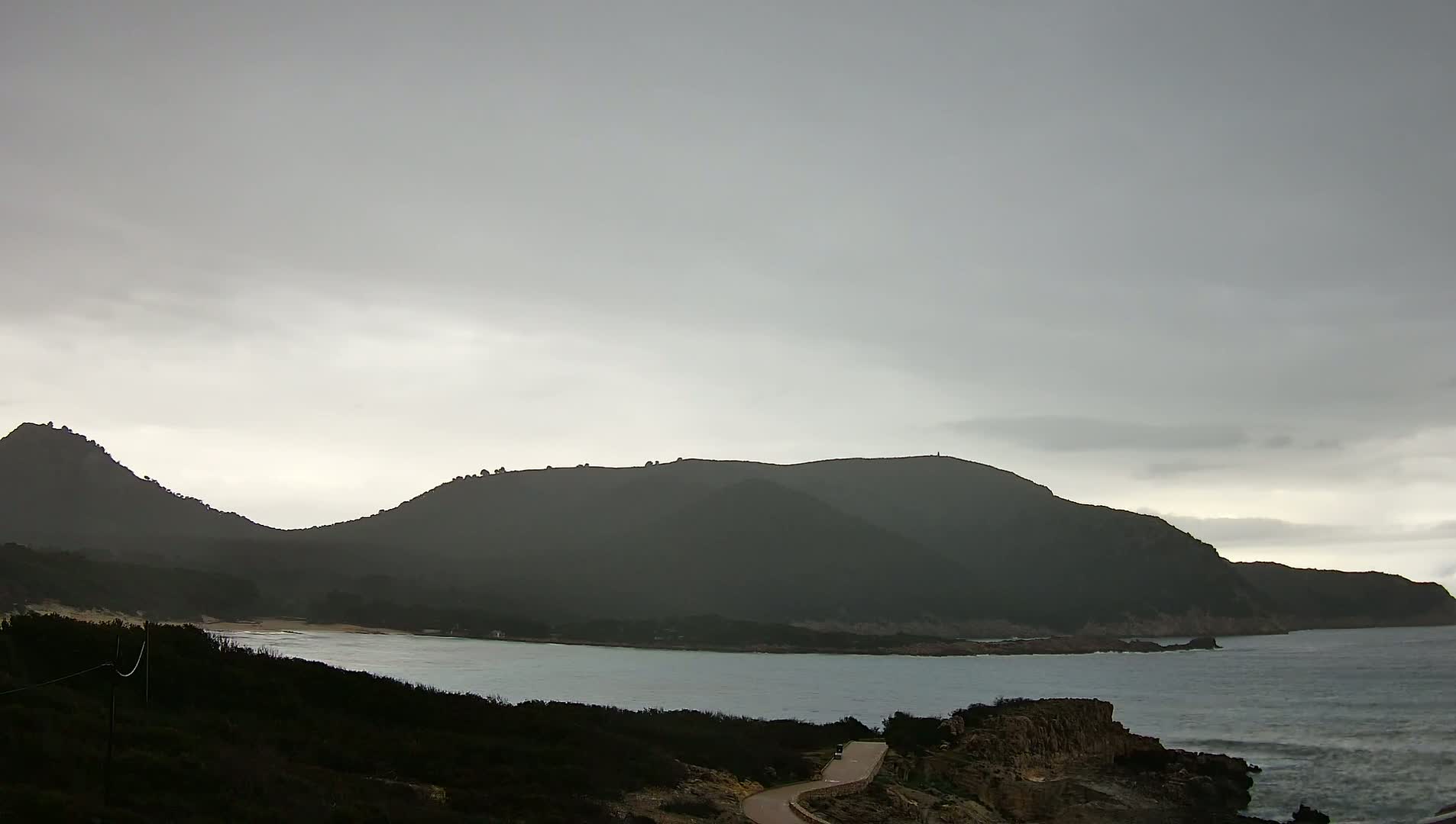 A calm bay is nestled between dark, rolling hills under an overcast sky.