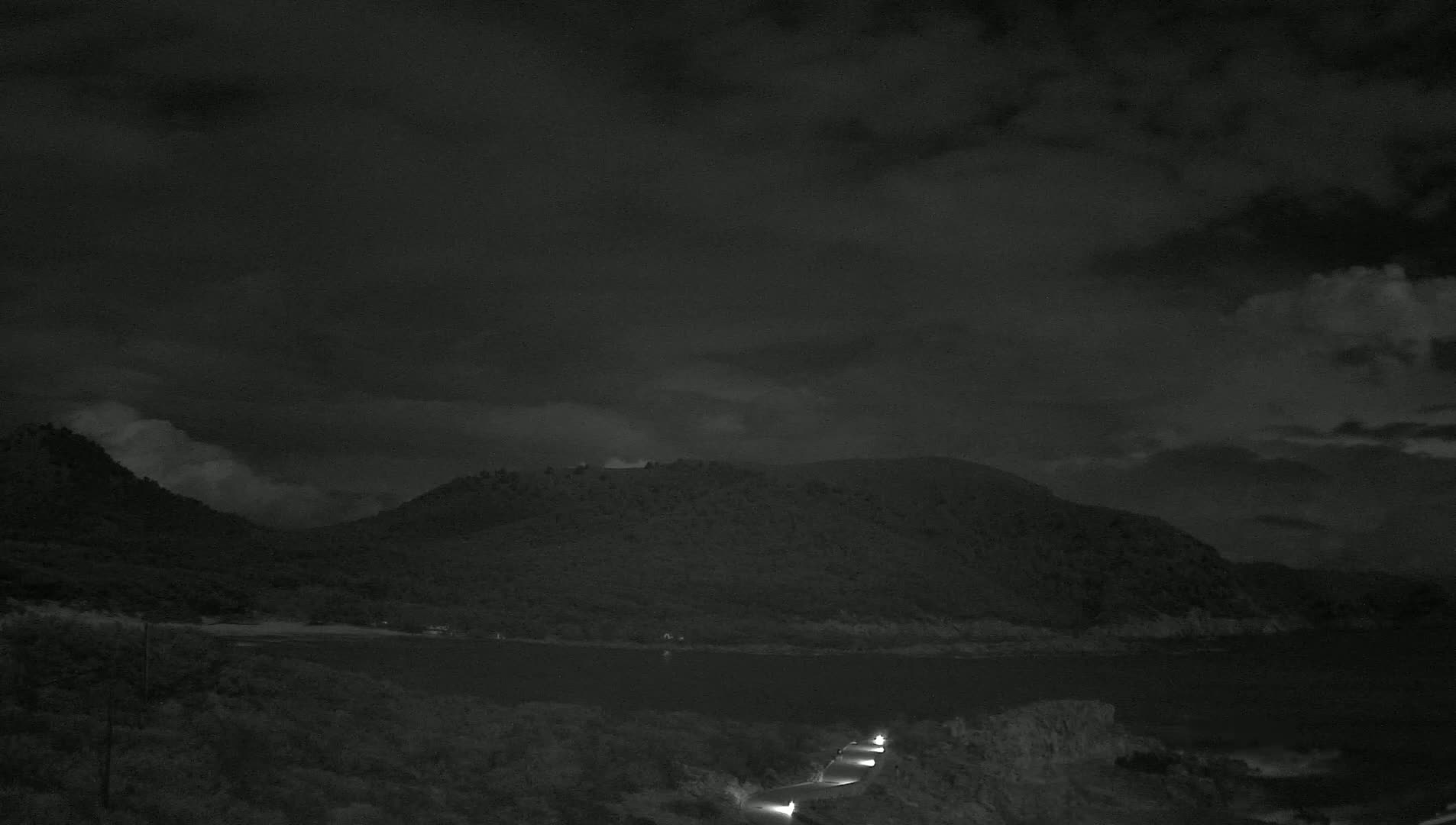 A nighttime view of a dark, cloudy sky over a calm body of water nestled between low, tree-covered hills, with a road and a few lights visible in the foreground.