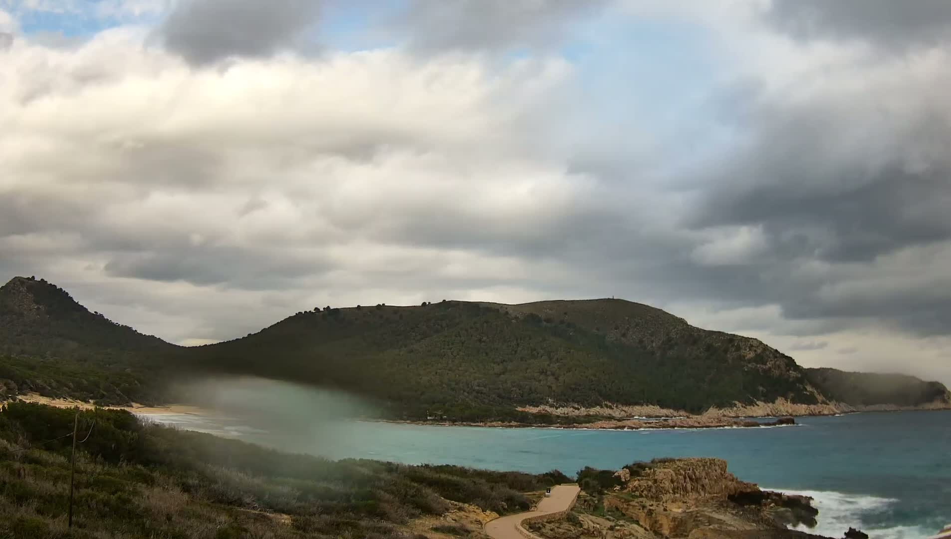 A tranquil bay is nestled between two verdant hills under a cloudy sky, with a hint of mist or spray near the water's edge.