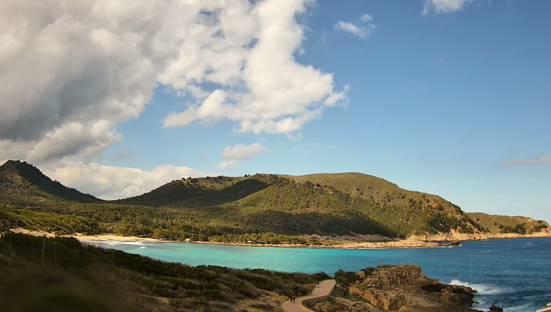 A partly cloudy sky overlooks a tranquil turquoise cove nestled between verdant hills and a rocky coastline.