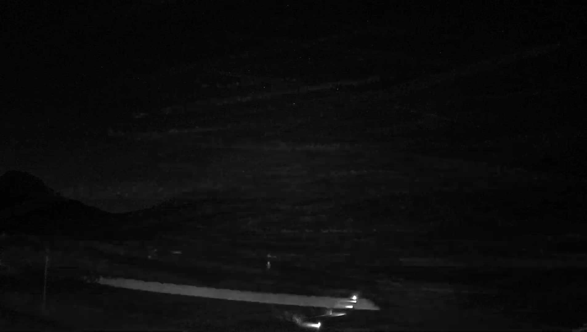 Under a partly cloudy night sky, headlights from vehicles are visible on a road below a dark mountain range.