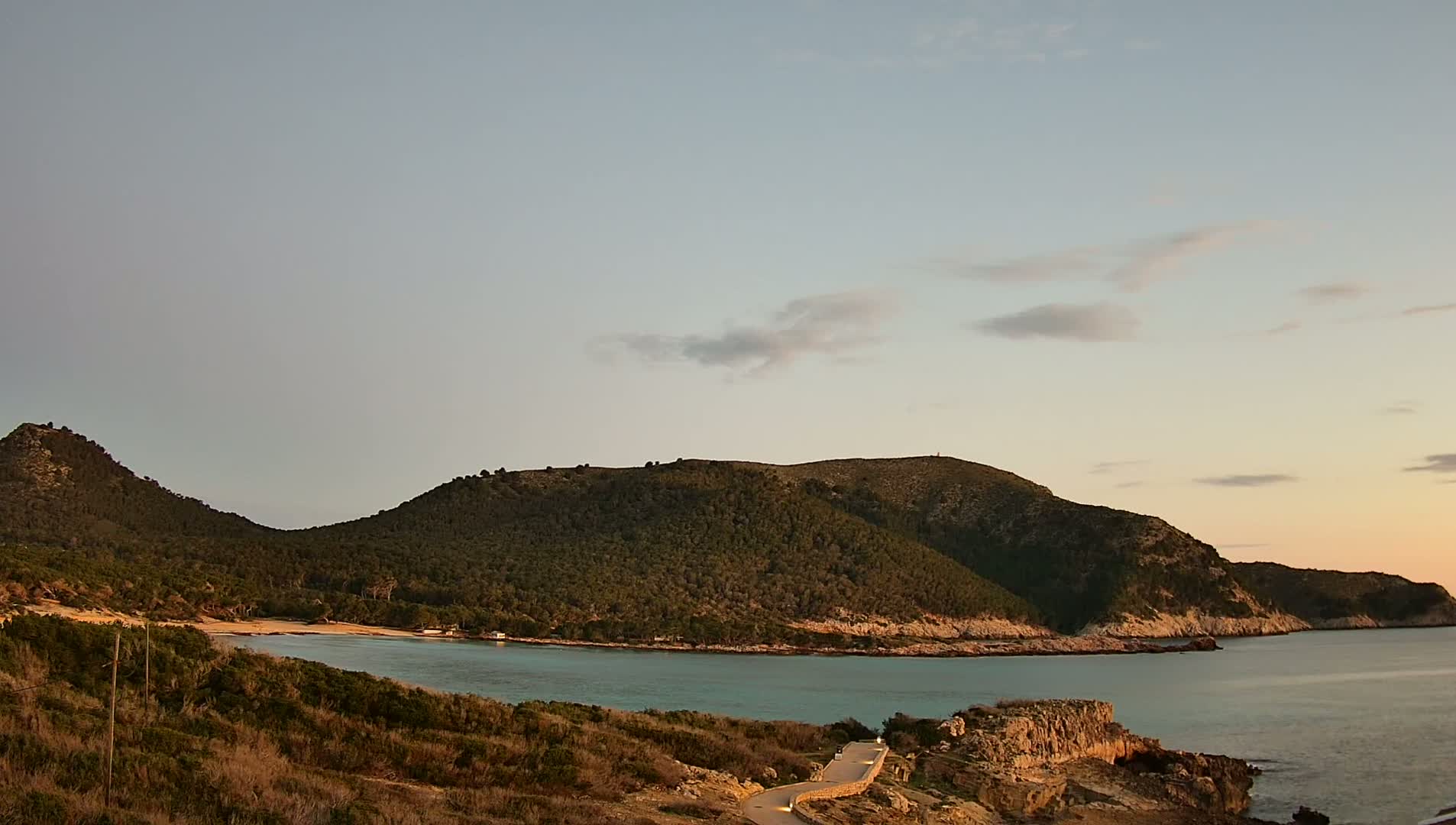 A tranquil cove with a small sandy beach is nestled between two verdant hills under a pale, mostly clear sky.