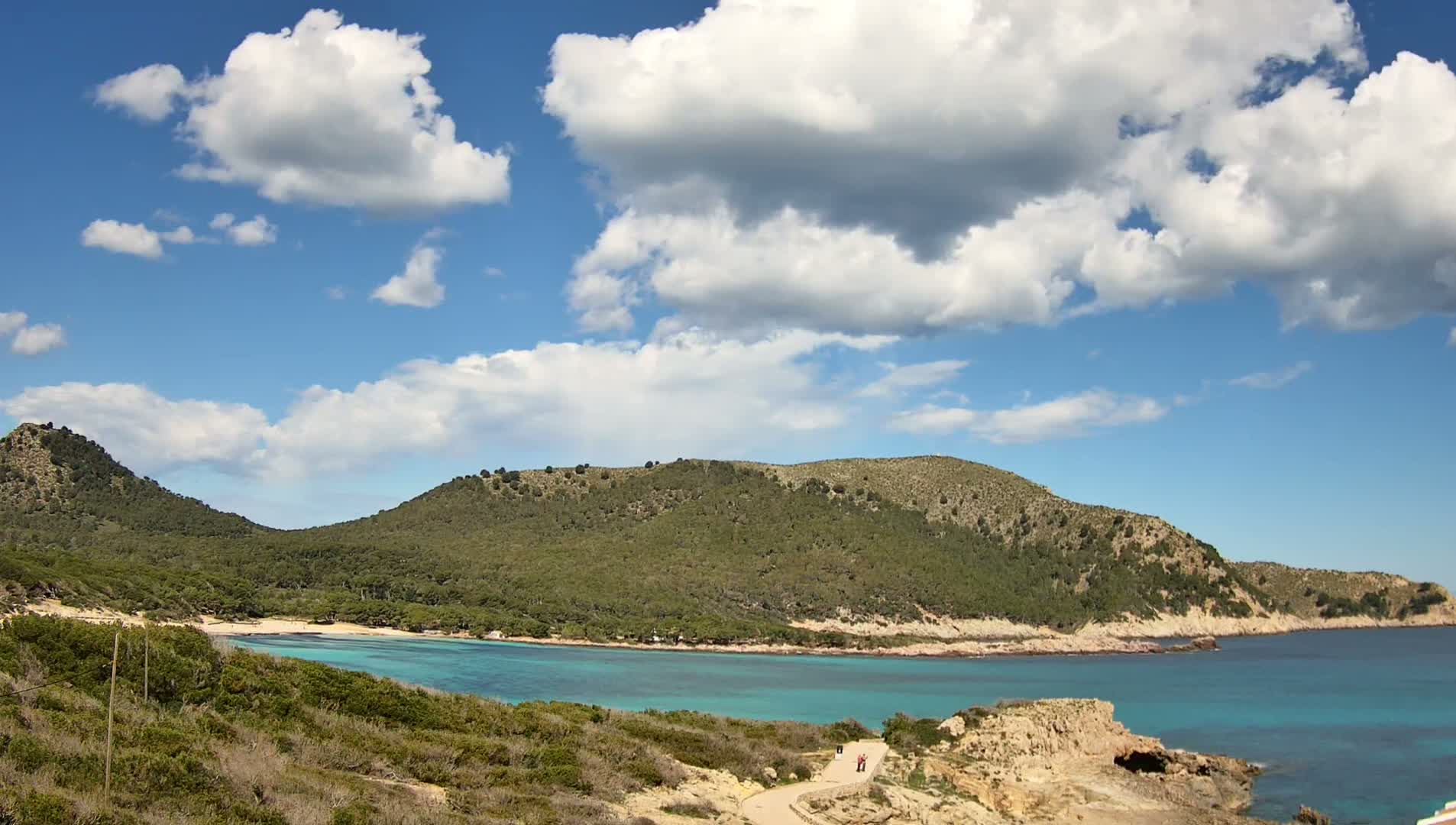 A secluded cove with turquoise water is nestled between green hills under a partly cloudy blue sky.