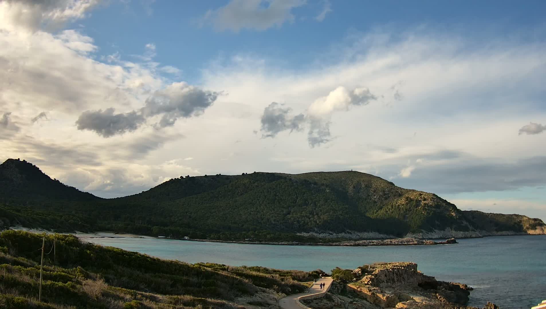 A partly cloudy sky overlooks a tranquil bay nestled between verdant hills, with a paved path winding along the coastline.