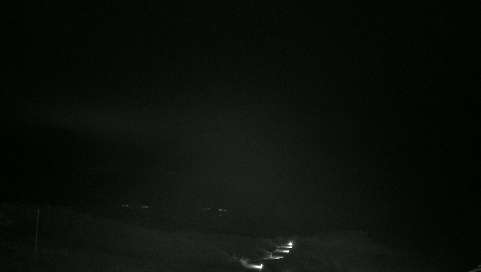 At night, a dark road with a few sets of headlights is visible against a dark, overcast sky.