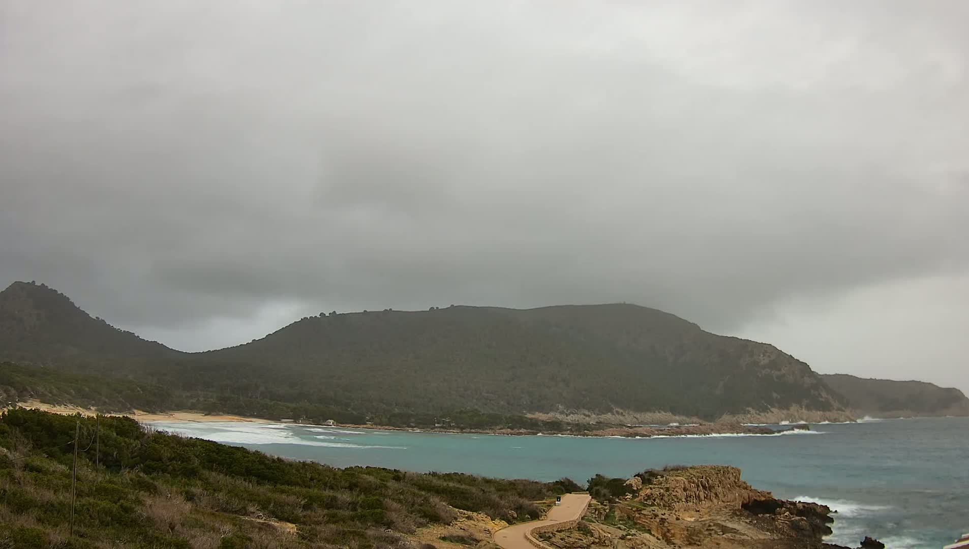 A secluded cove with a sandy beach is nestled between dark green hills under a heavily overcast sky, with choppy waves visible in the ocean.