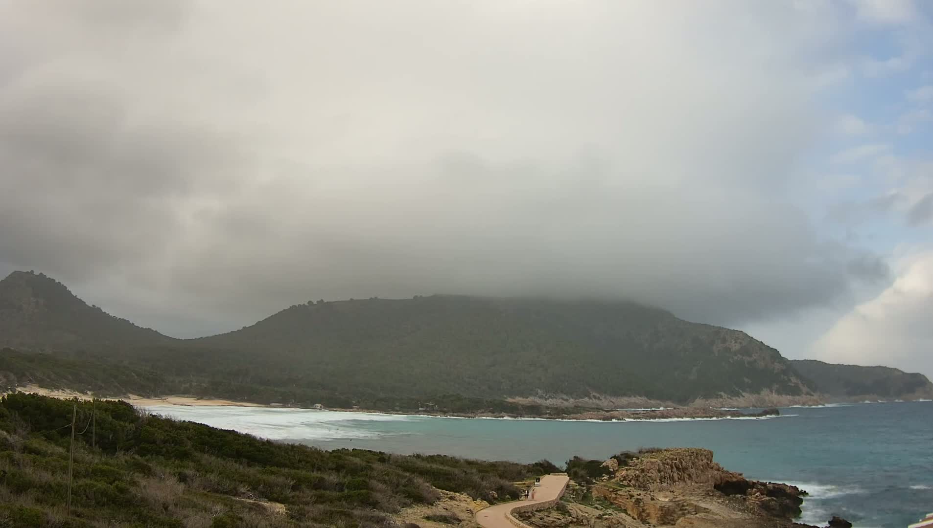A coastal scene shows a partly cloudy sky over a green, mountainous coastline with a sandy beach and turquoise water.