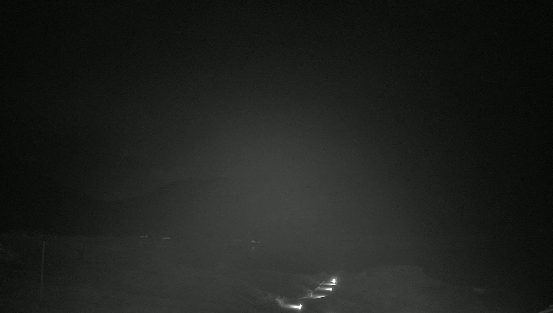 Faintly lit vehicles are visible on a dark road at night under foggy or hazy conditions.