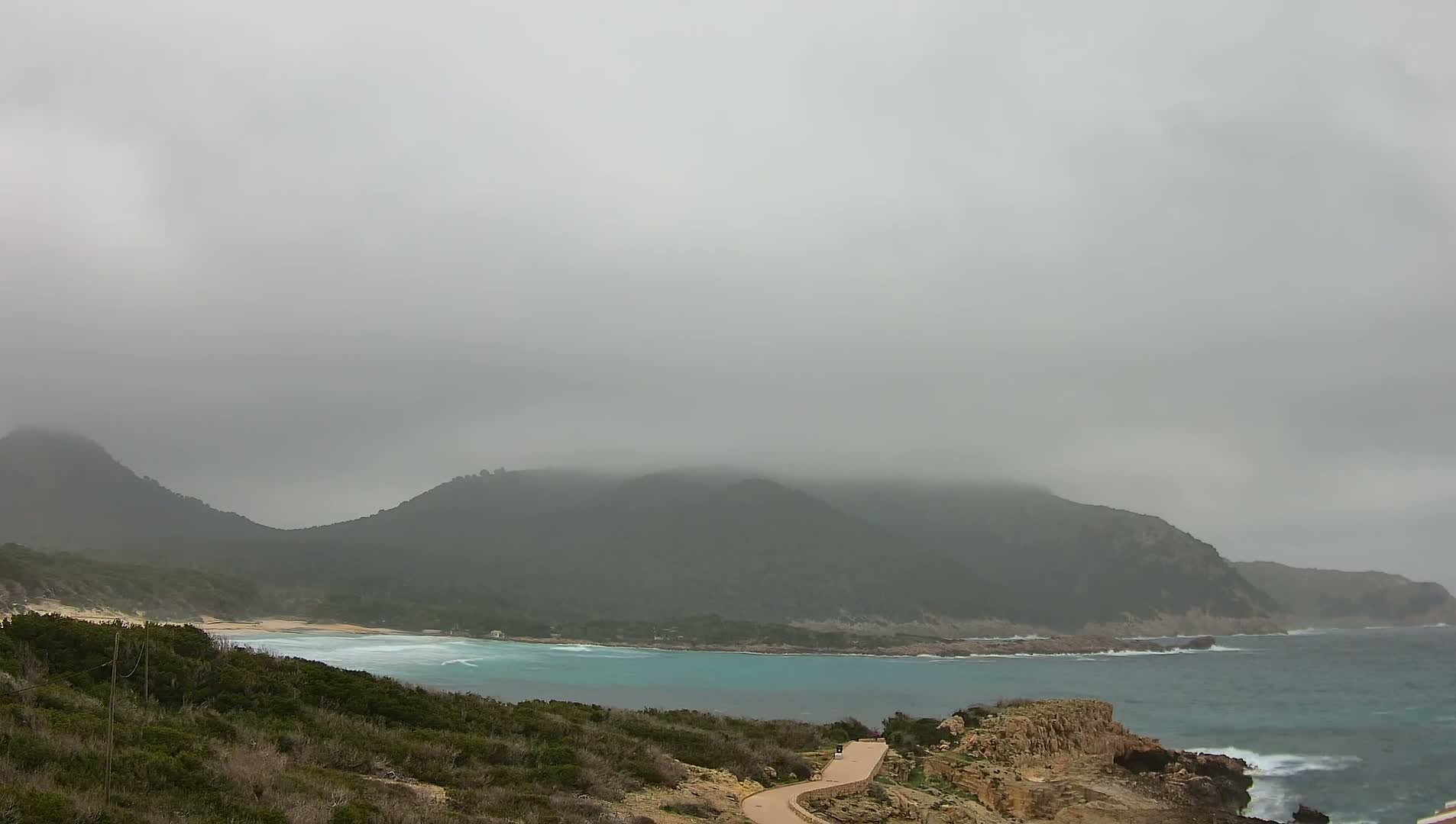 A foggy coastal scene shows a secluded cove with turquoise water, a sandy beach, low-lying vegetation, and rocky hills shrouded in mist.