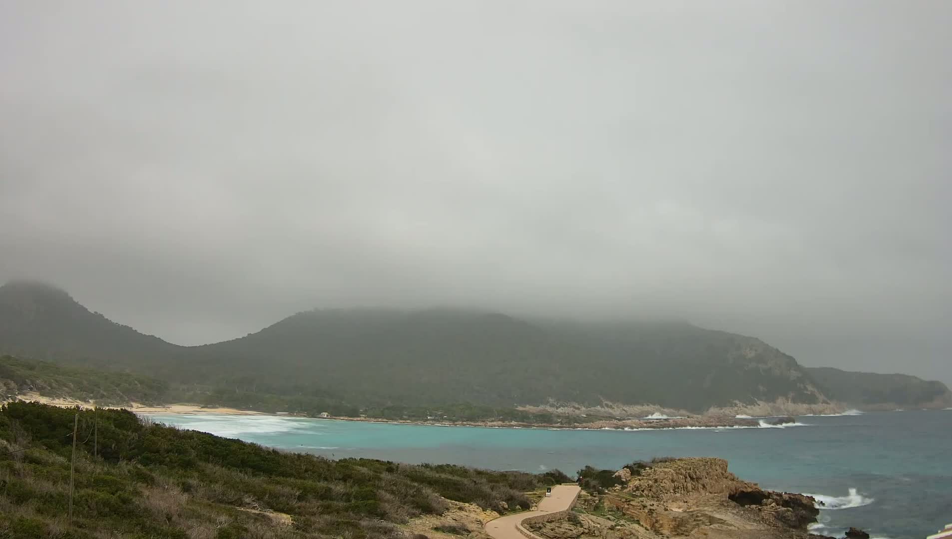 A fog-shrouded mountain overlooks a calm, turquoise bay with a sandy beach and a paved path leading down to the shore.