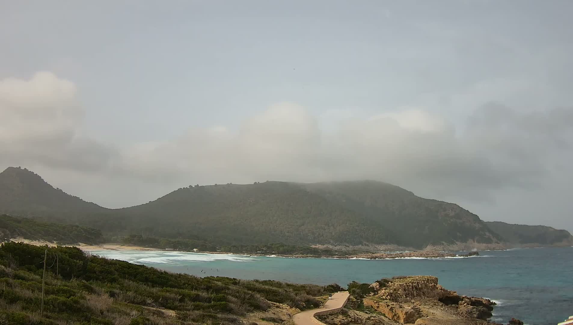 A hazy, overcast day reveals a secluded cove with turquoise water lapping a sandy beach, nestled between verdant hills under a mostly cloudy sky.