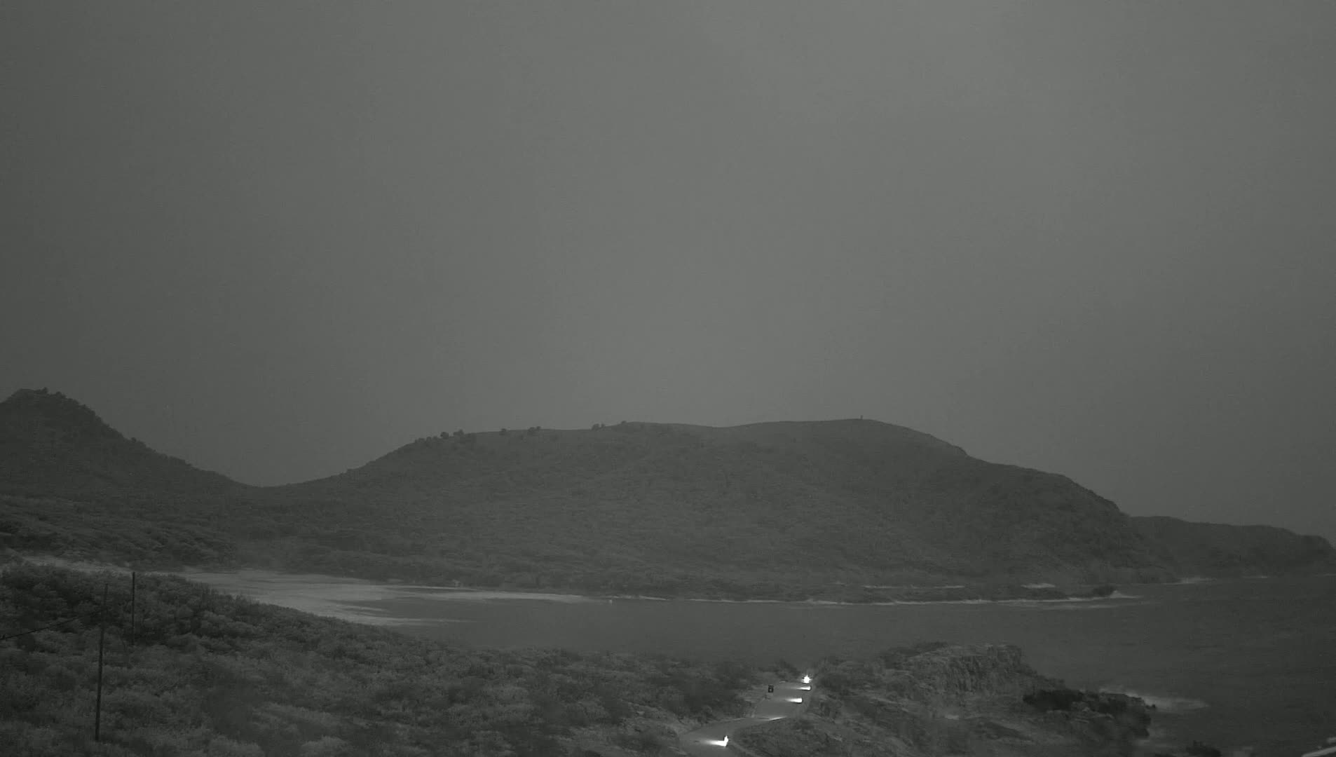 A hazy, dark gray sky hangs over a hilly, coastal landscape at dusk or dawn, with a road winding along the shoreline where a few vehicle headlights are visible.