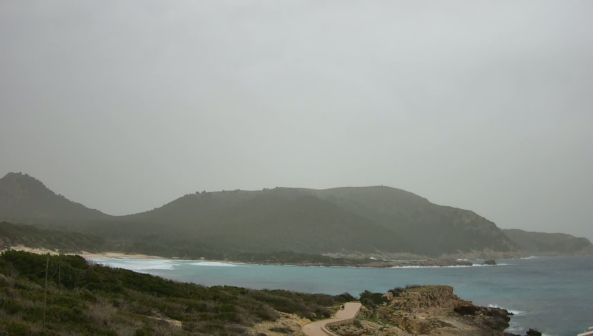 A hazy coastal scene shows a curving bay with light-blue water lapping a sandy beach, backed by low, green hills under a gray sky.