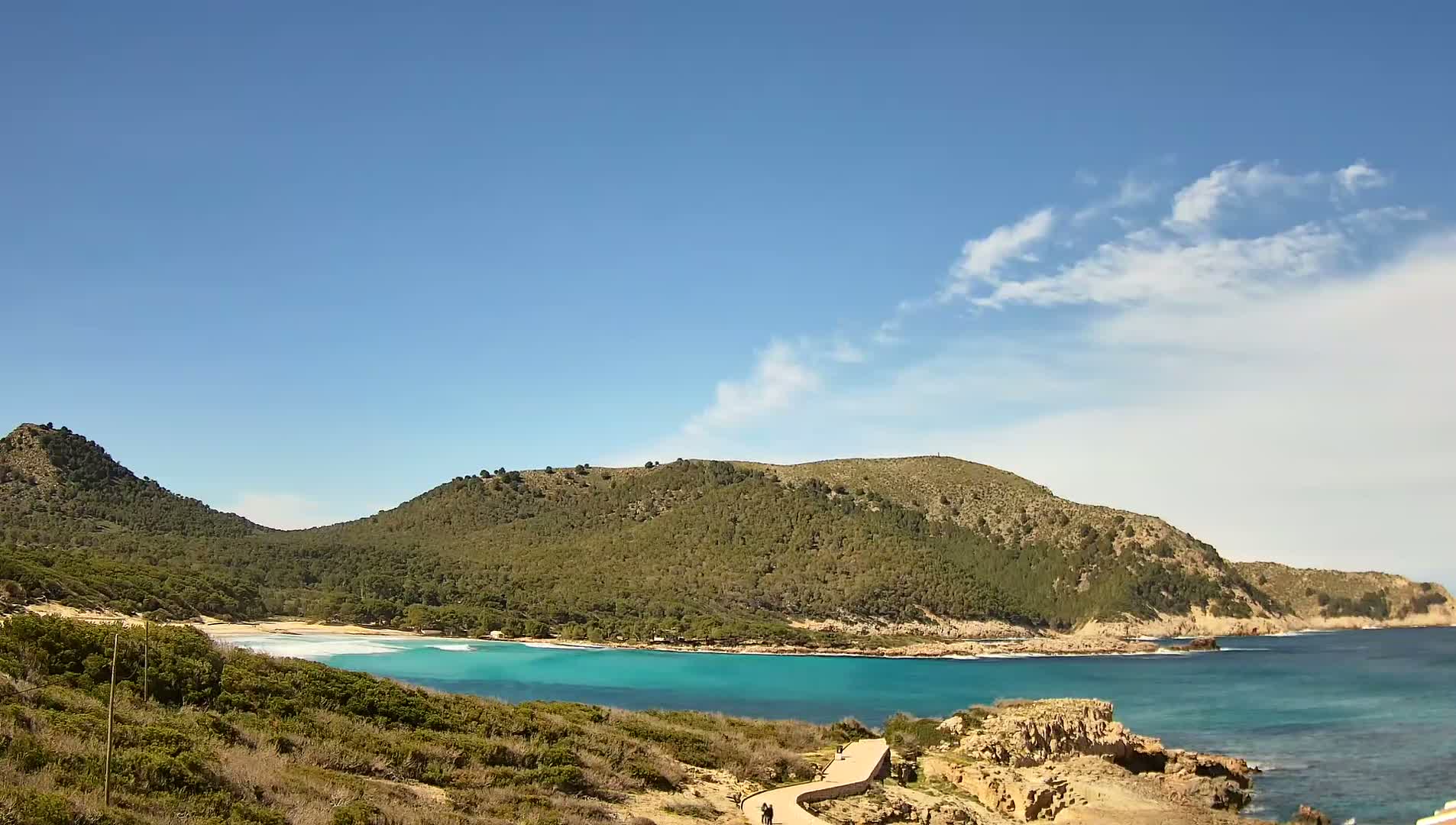 A secluded cove with turquoise water is nestled between two verdant hills under a mostly clear sky with a few scattered clouds.