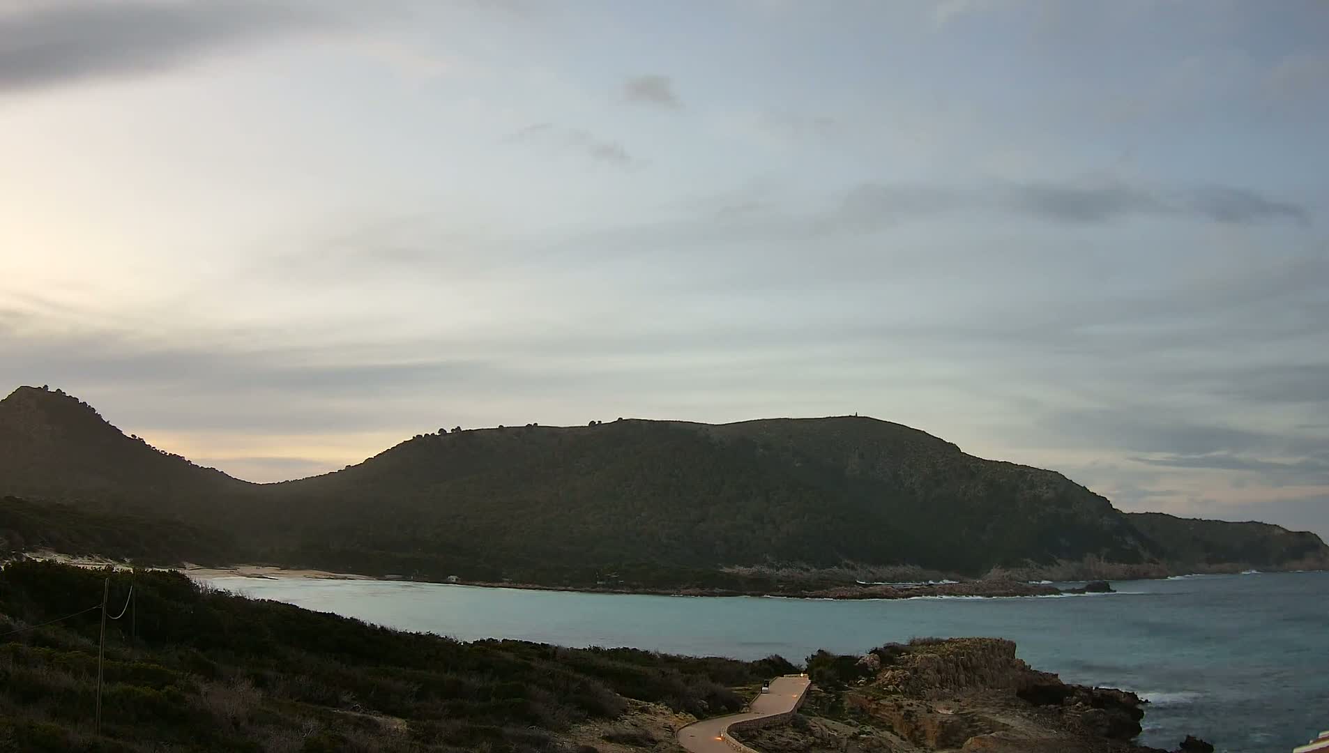 A winding road leads down to a calm, light-blue bay nestled between two dark, verdant hills under a cloudy sky.