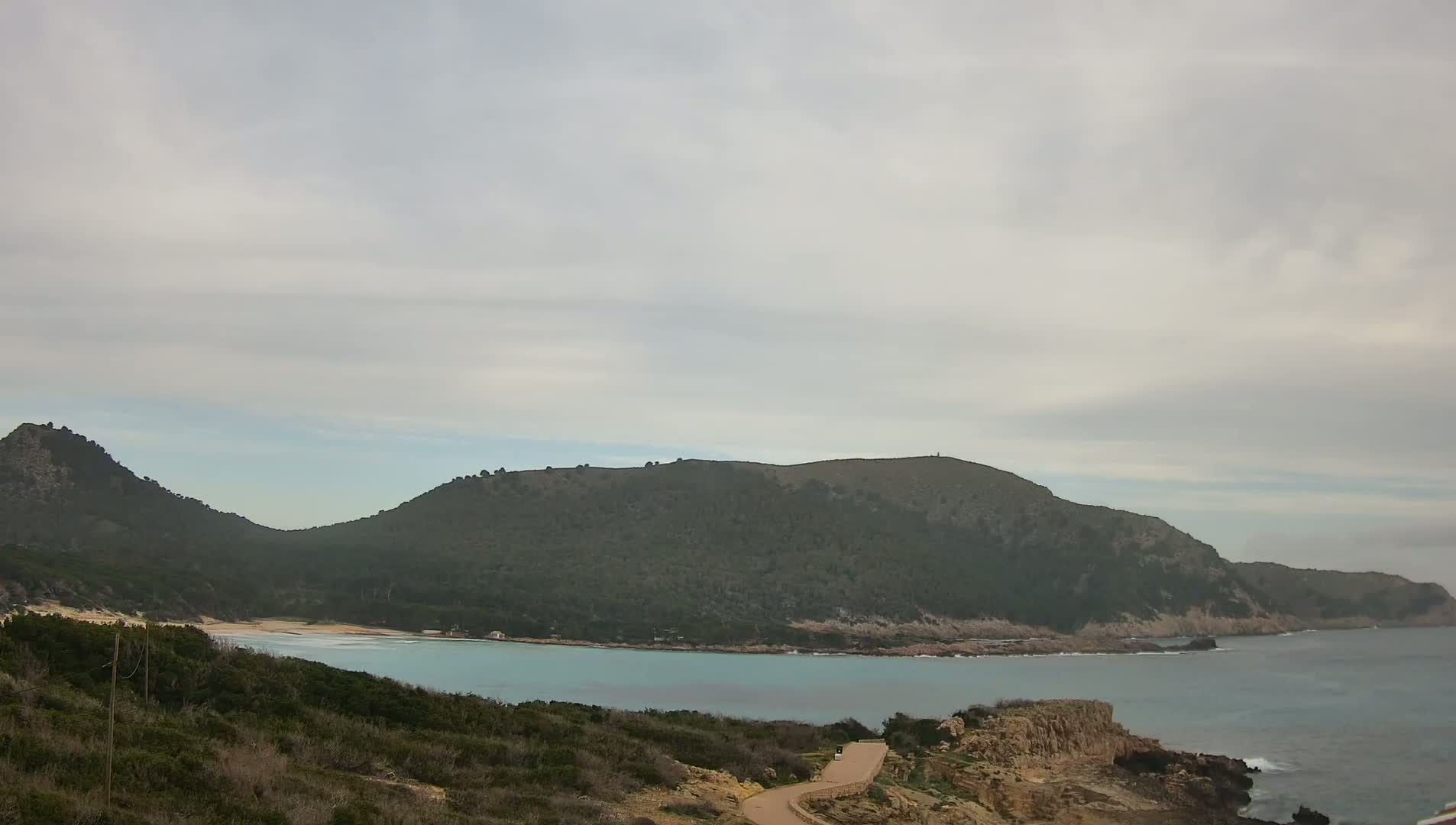 A secluded cove with a sandy beach is nestled between two verdant hills under a cloudy sky.