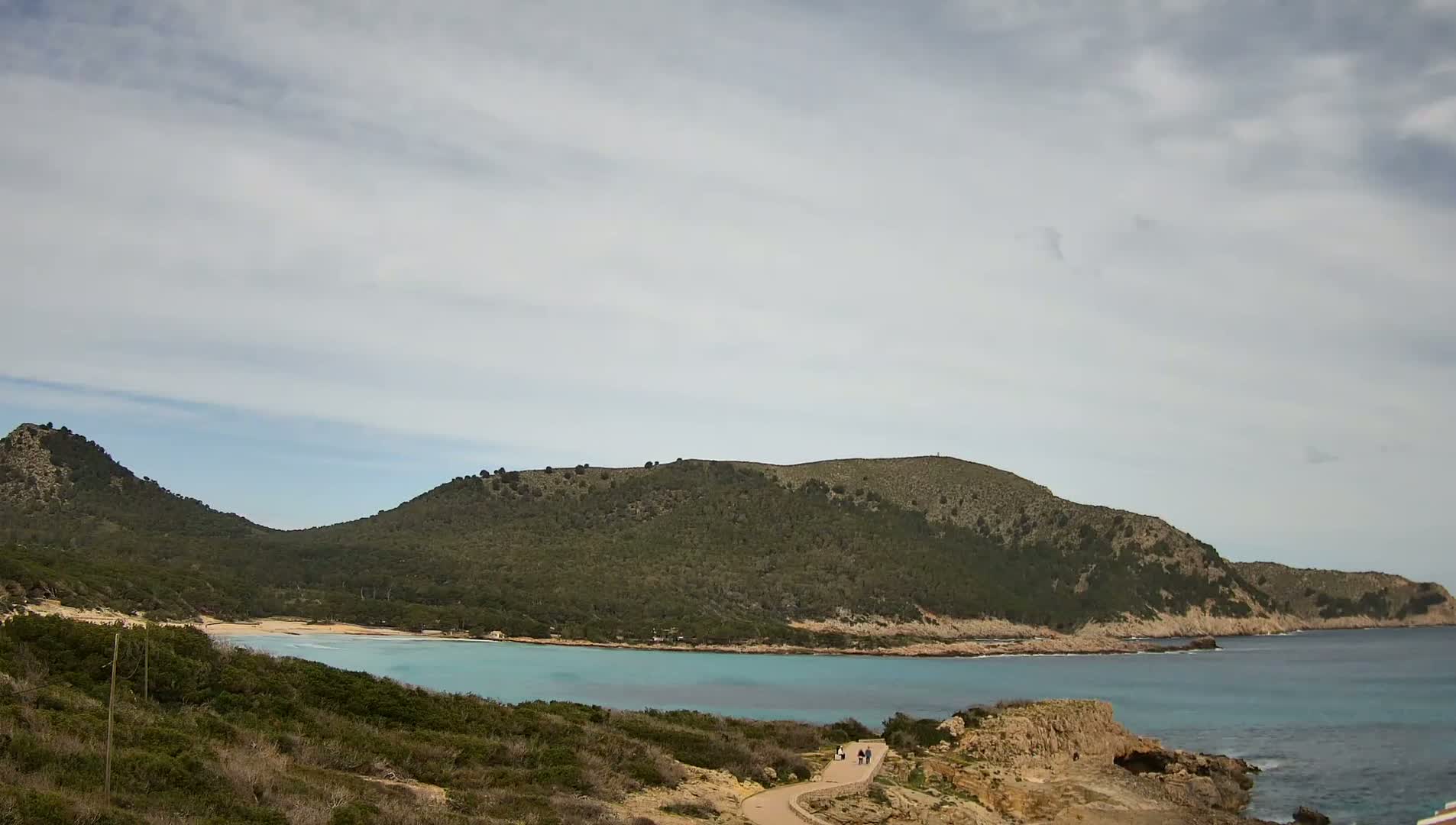 A partly cloudy sky overlooks a tranquil cove with a sandy beach nestled between two verdant hills, where a few people stroll along a path.