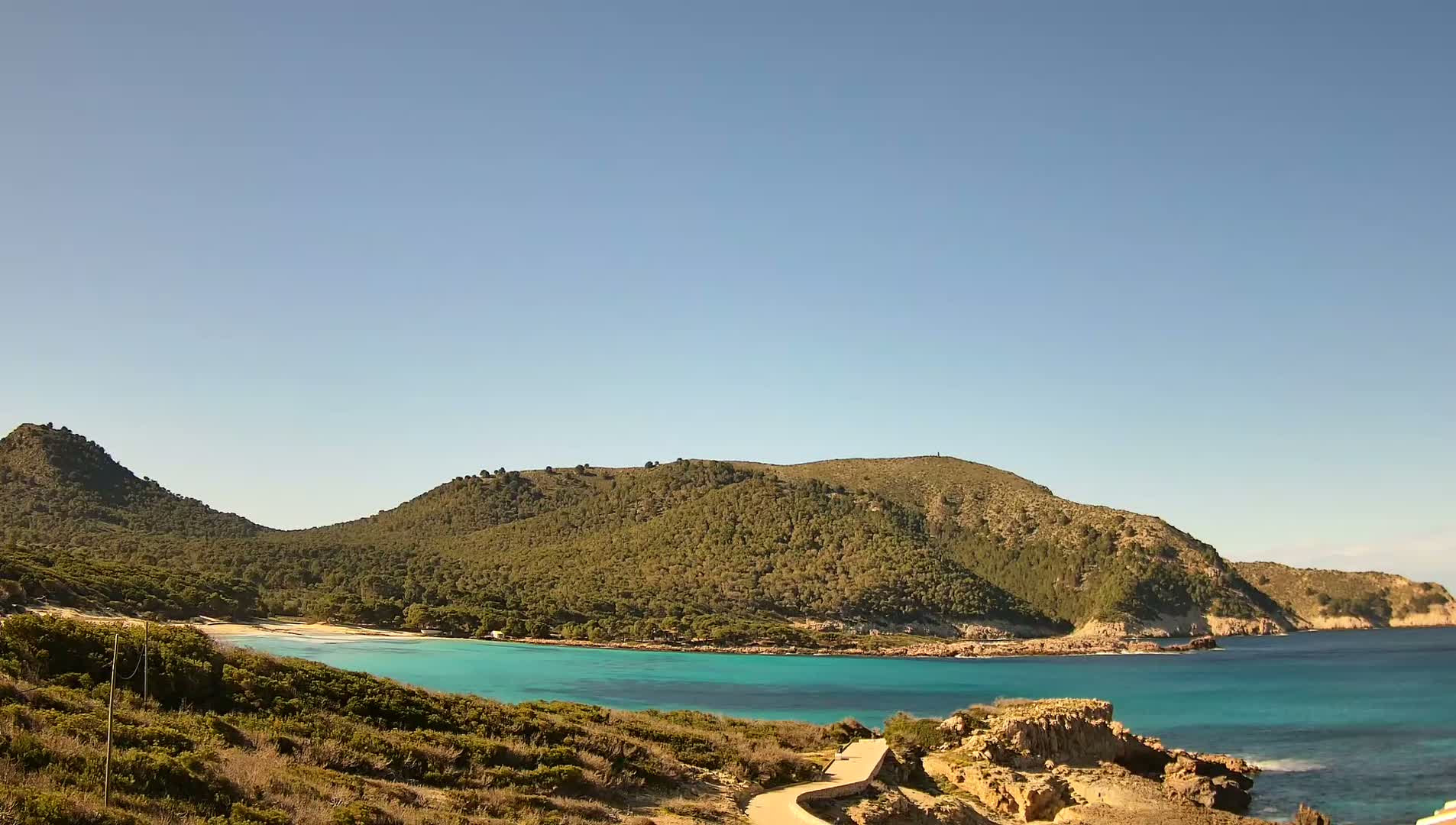 A secluded cove with a small, sandy beach and turquoise water is nestled between two verdant hills under a clear, sunny sky.