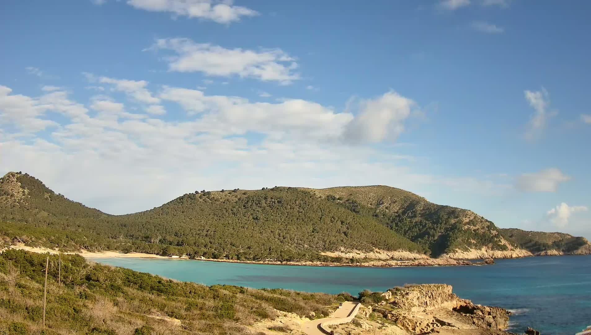 A partly cloudy sky overlooks a secluded cove with a small sandy beach nestled between two verdant hillsides.
