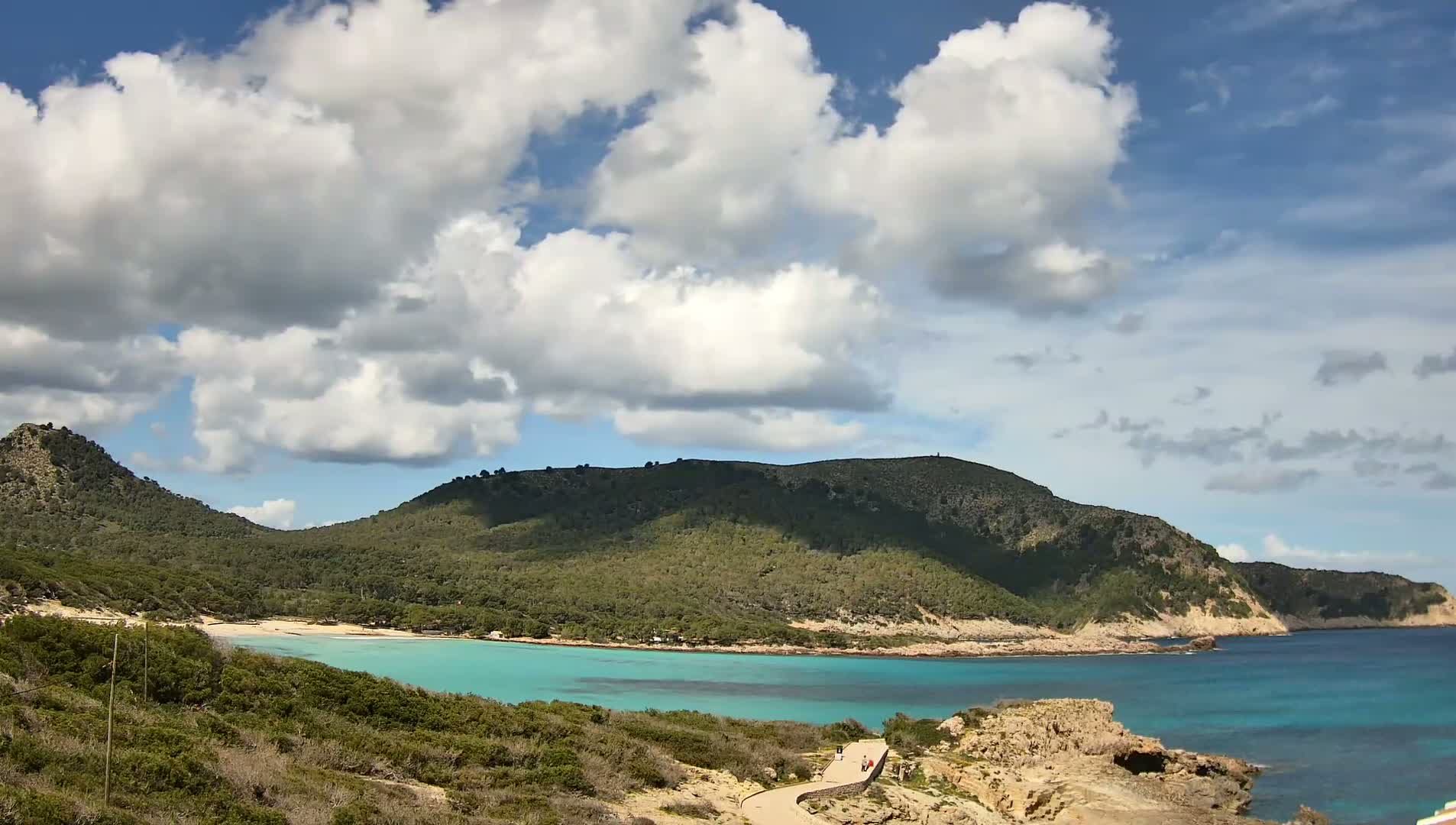 A partly cloudy sky overlooks a tranquil turquoise bay nestled between lush green hills, with a sandy beach visible at the bay's edge.