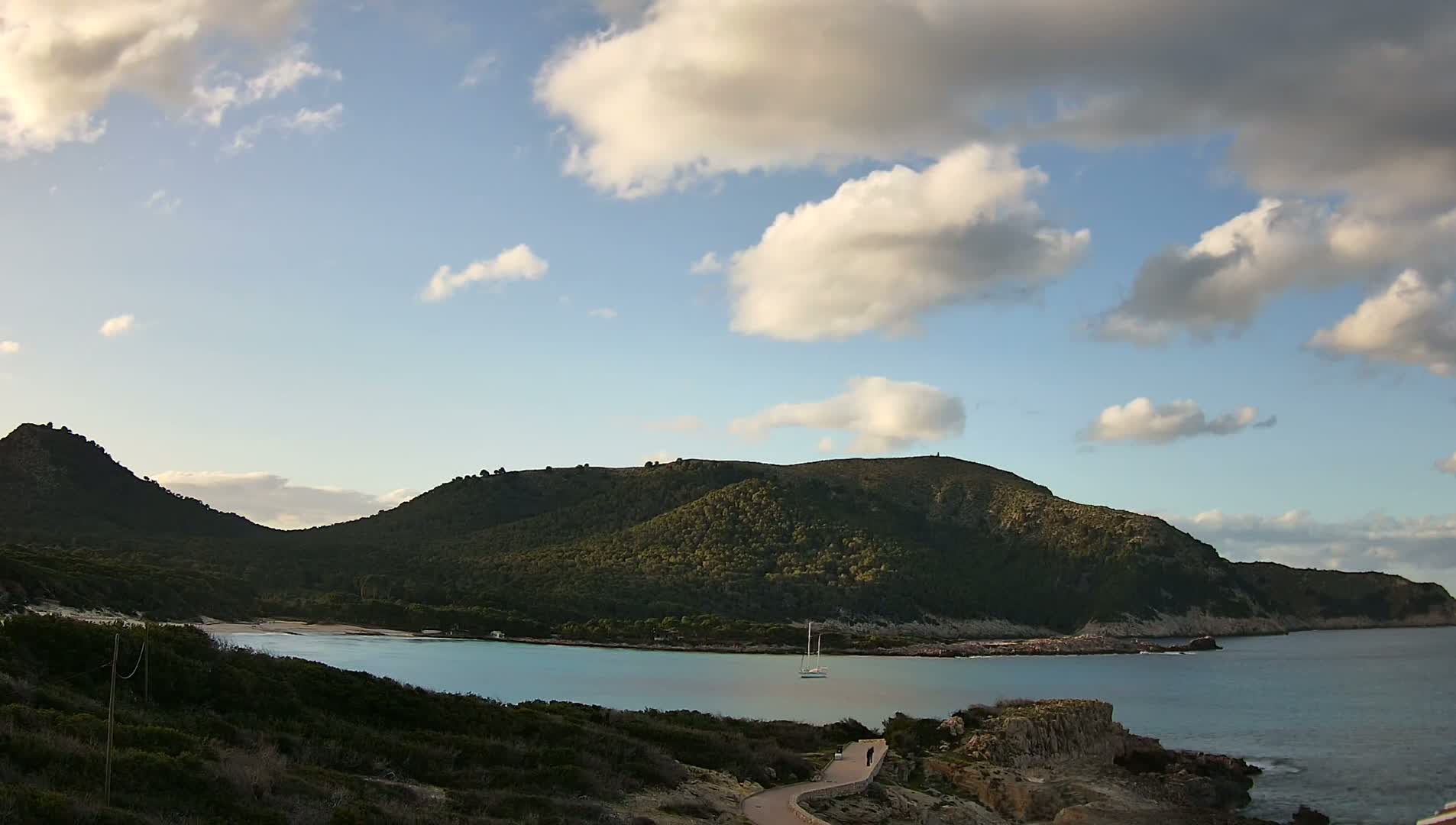 A partly cloudy sky overlooks a tranquil bay nestled between two verdant hills, with a lone sailboat at anchor.