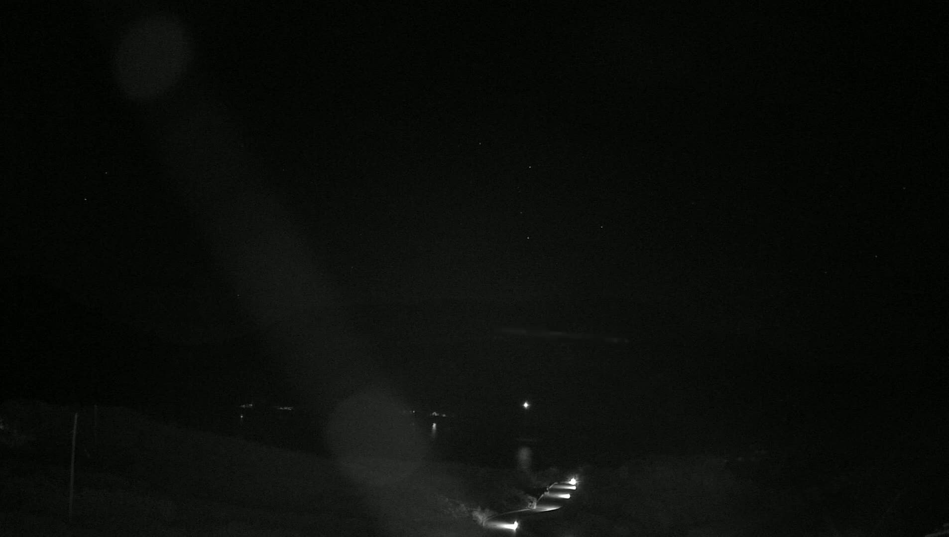 A nighttime, low-light view of a road with a few headlights visible, under a dark, clear sky.