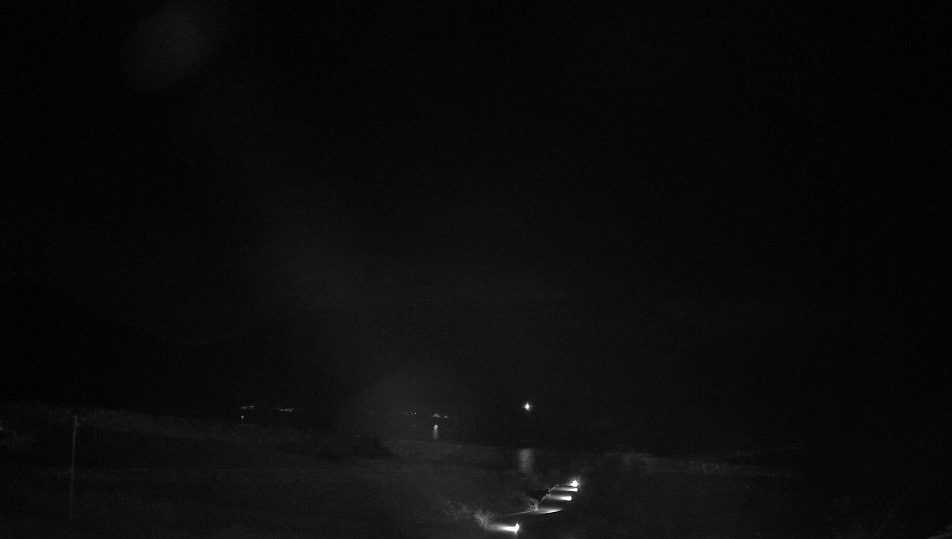 At night, under a dark sky, a road with a few vehicles is visible, with some faint lights in the distance.