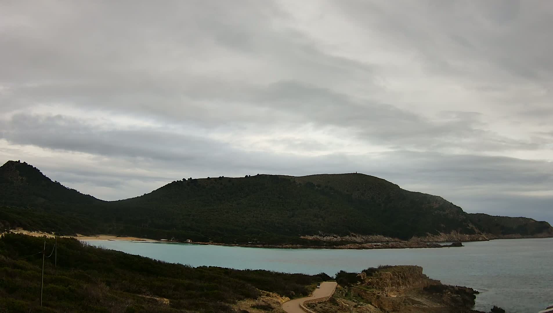 A secluded cove with a light teal bay is nestled between two verdant hills under an overcast sky.