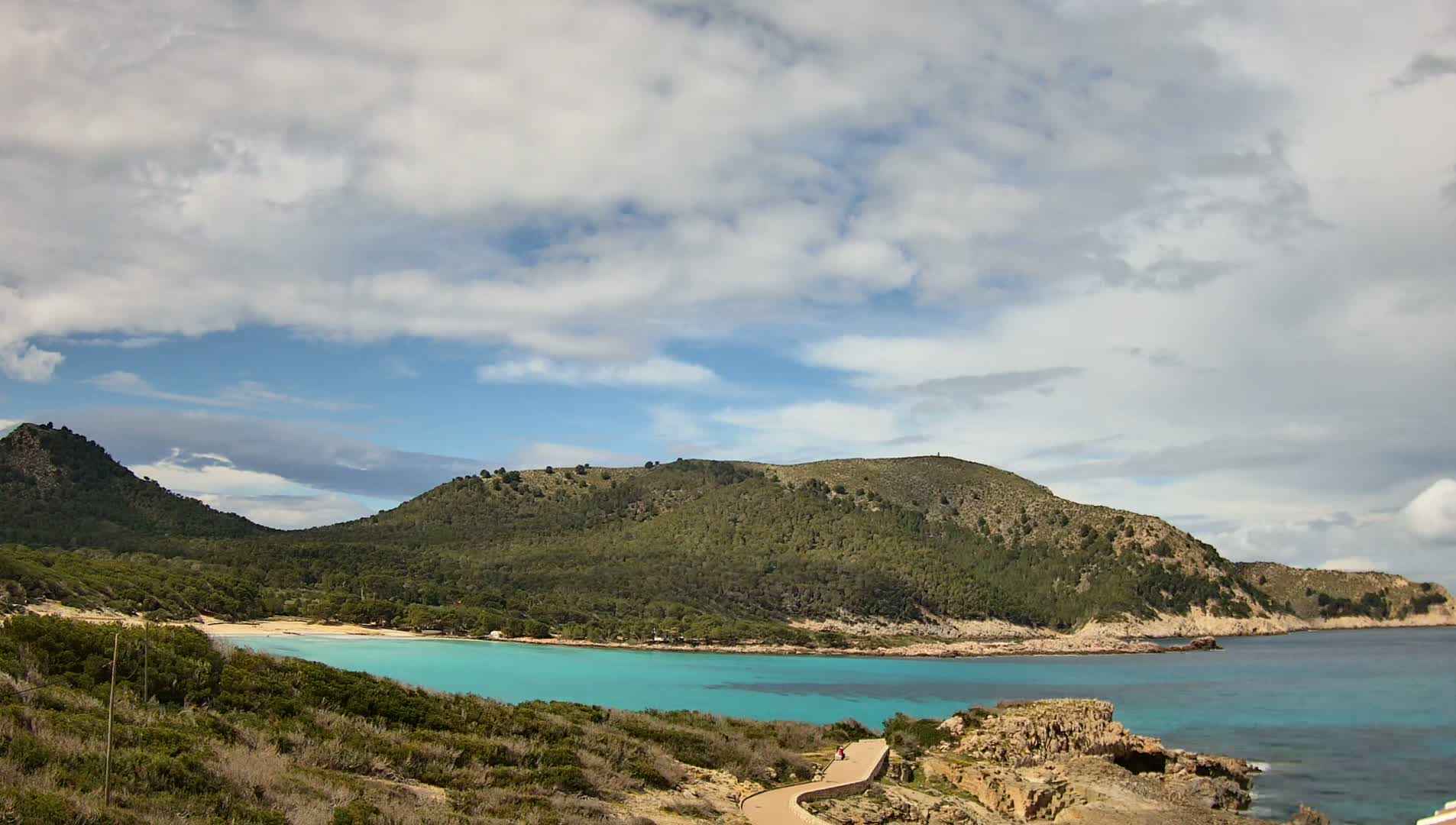A partly cloudy sky overlooks a tranquil turquoise cove nestled between verdant hills, with a paved pathway winding down towards the beach.
