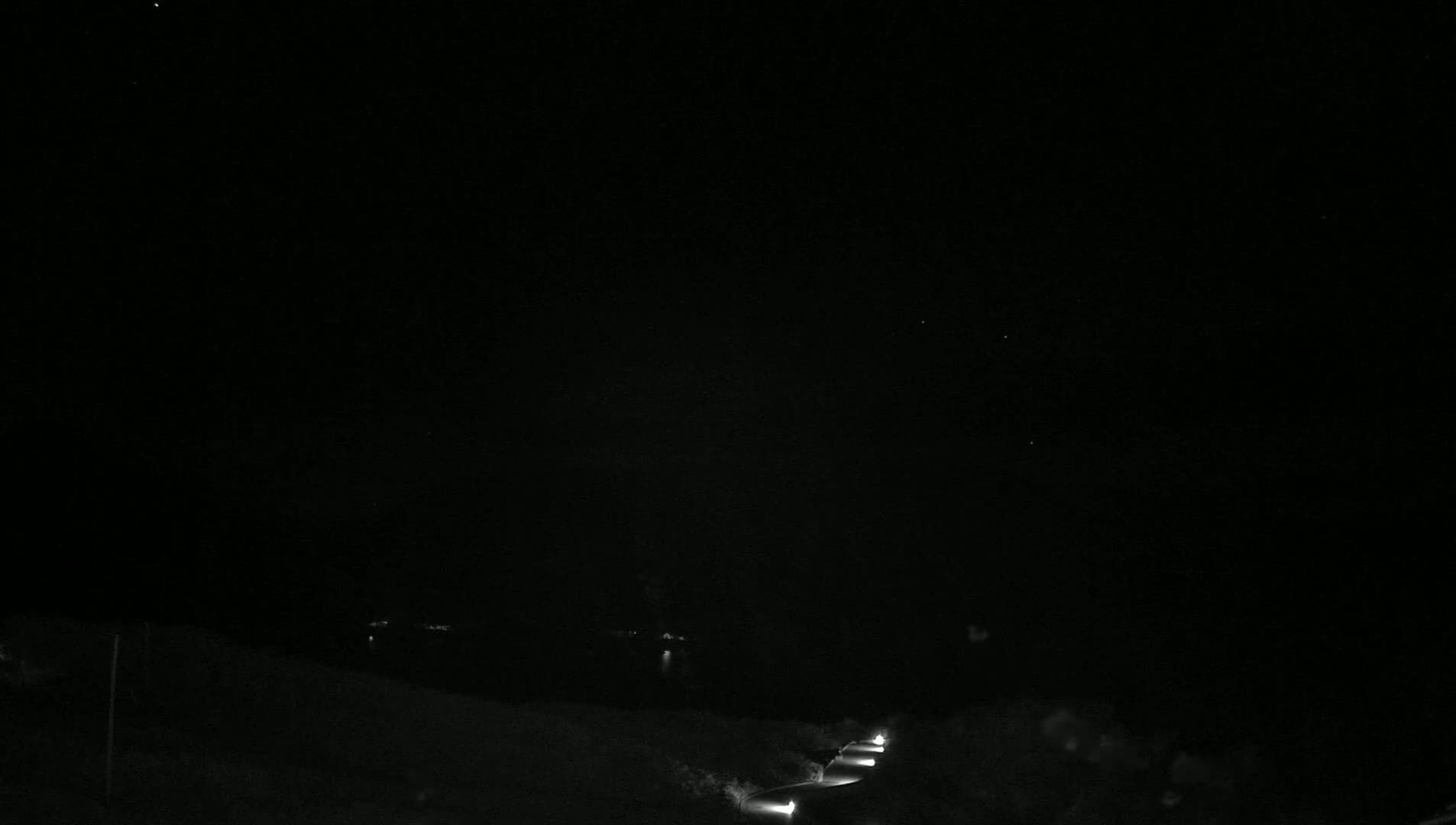 A dark nighttime scene shows a road with a few headlights visible in the distance, leading toward a body of water under a clear, starlit sky.