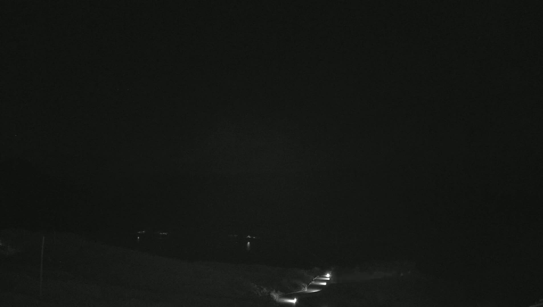 A nighttime, dark scene shows a road curving through a landscape with a few distant lights visible.