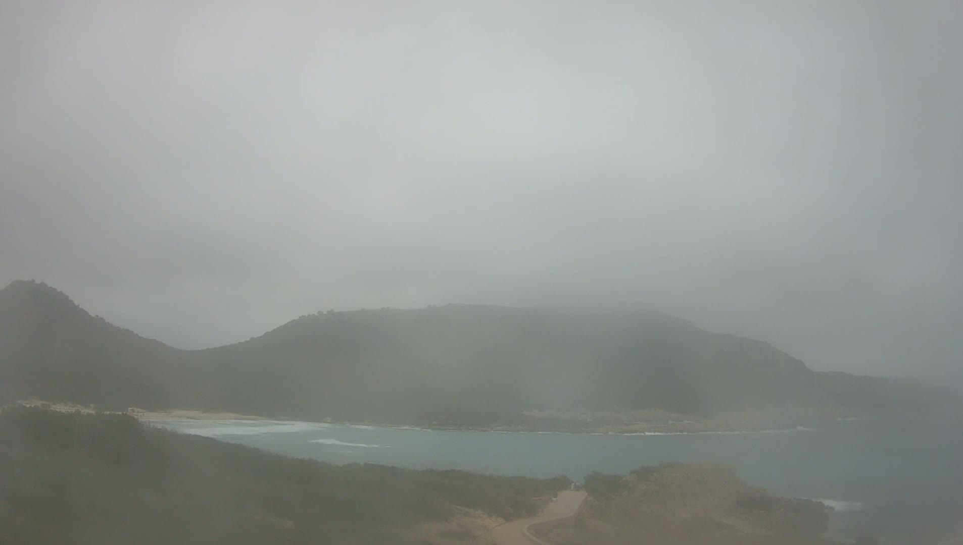A foggy coastal scene shows a body of water nestled between two hills.