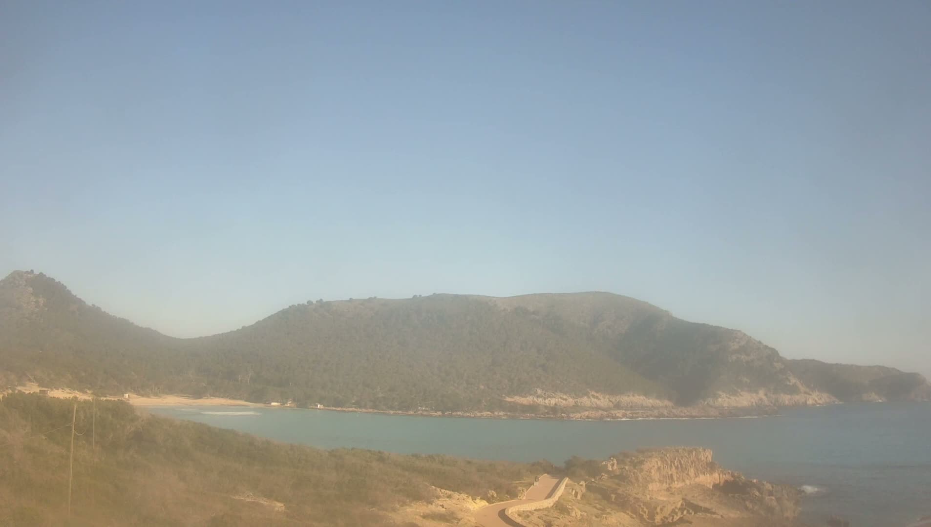 A hazy, sunny day reveals a secluded cove nestled between two verdant hillsides, with a calm sea stretching to the horizon.